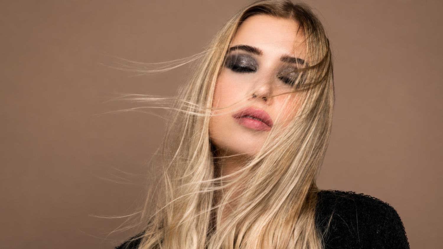 Blonde woman with smoky eye makeup and windswept hair posing against a warm beige background.
