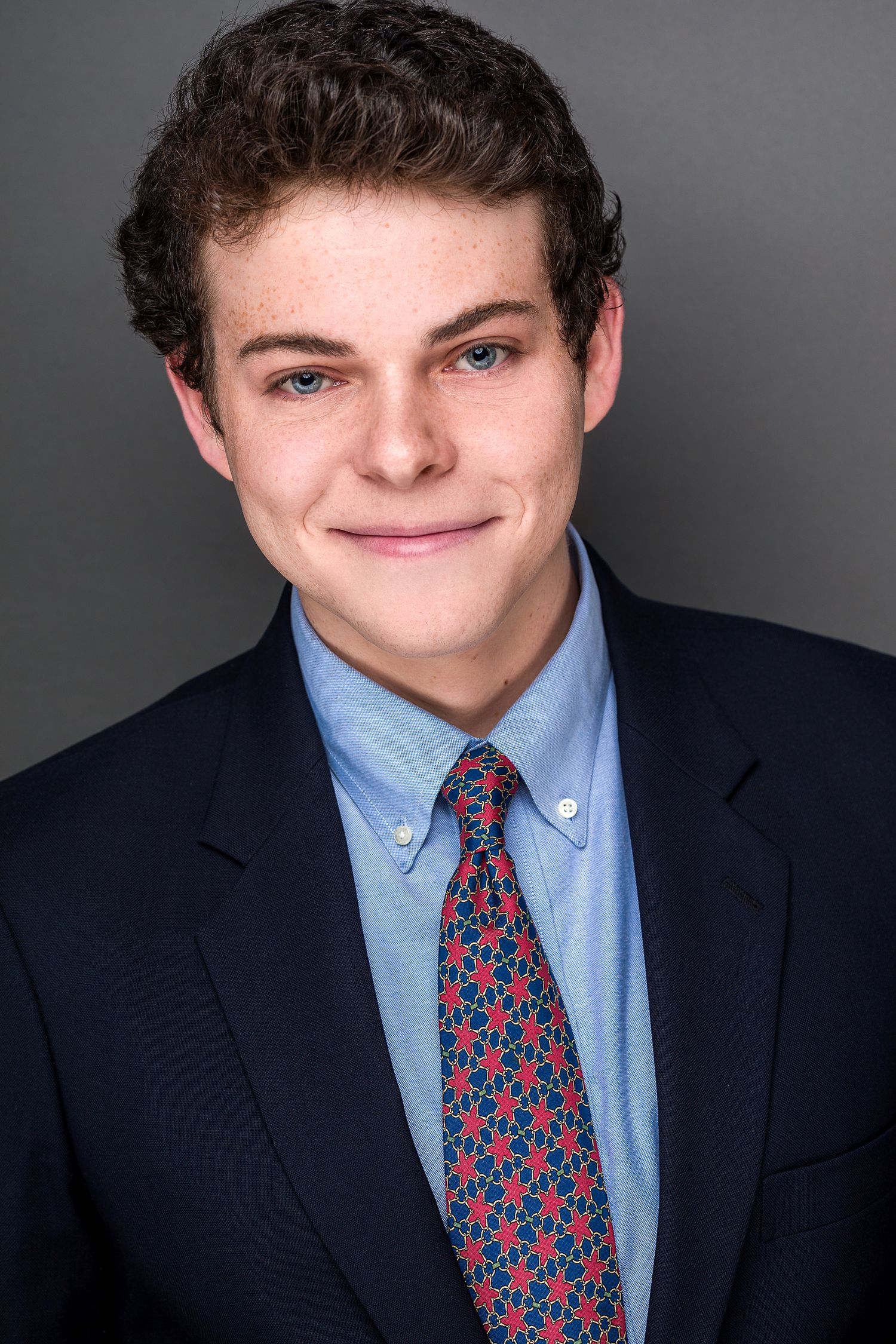Professional headshot of a young man in a navy suit with blue shirt and red patterned tie, smiling warmly.