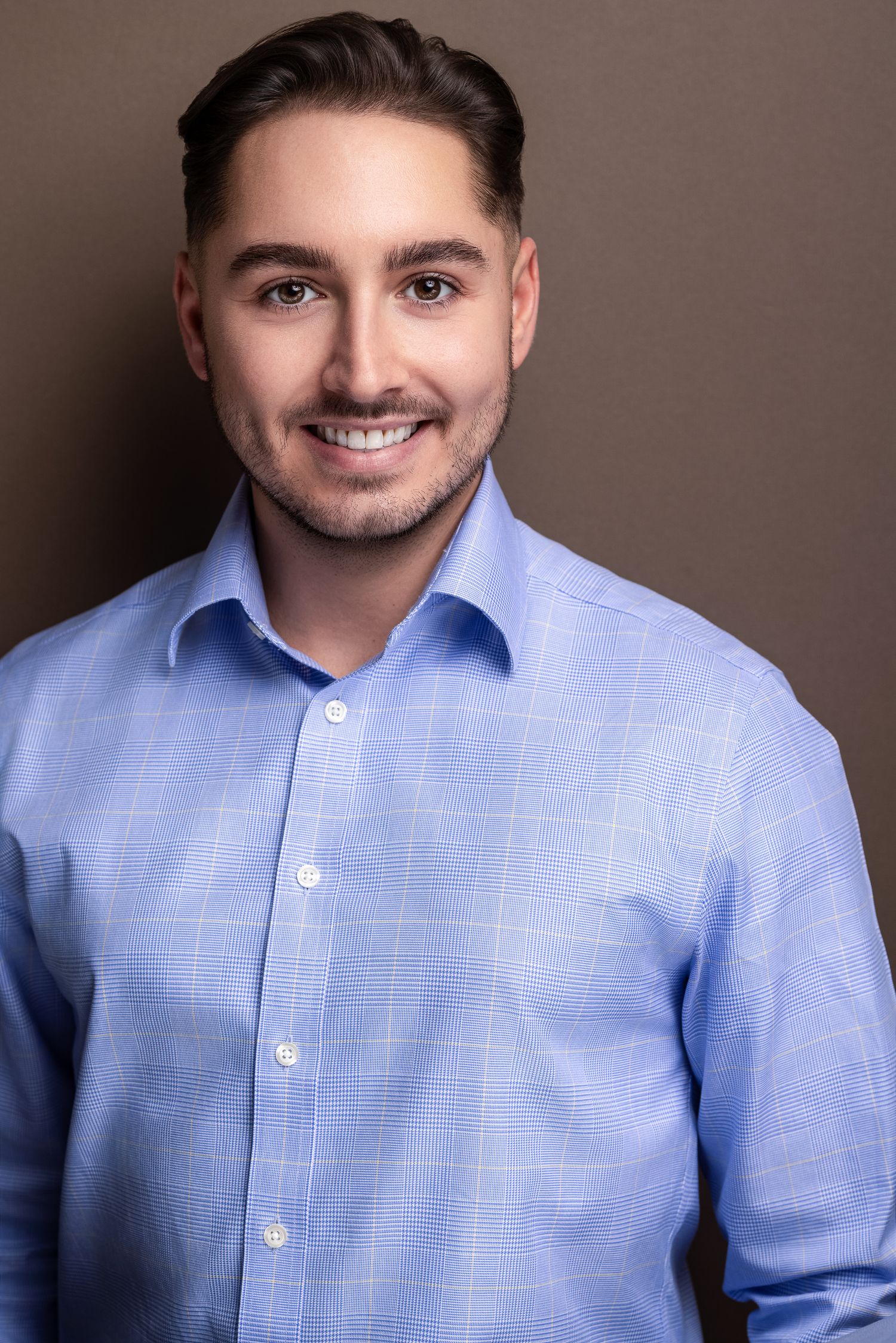Professional headshot of a smiling young man with dark hair wearing a light blue dress shirt against a gray background.