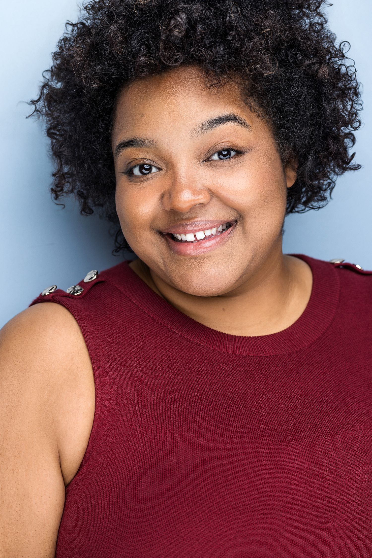 Smiling young woman with curly hair wearing a red sleeveless top against a light blue background.