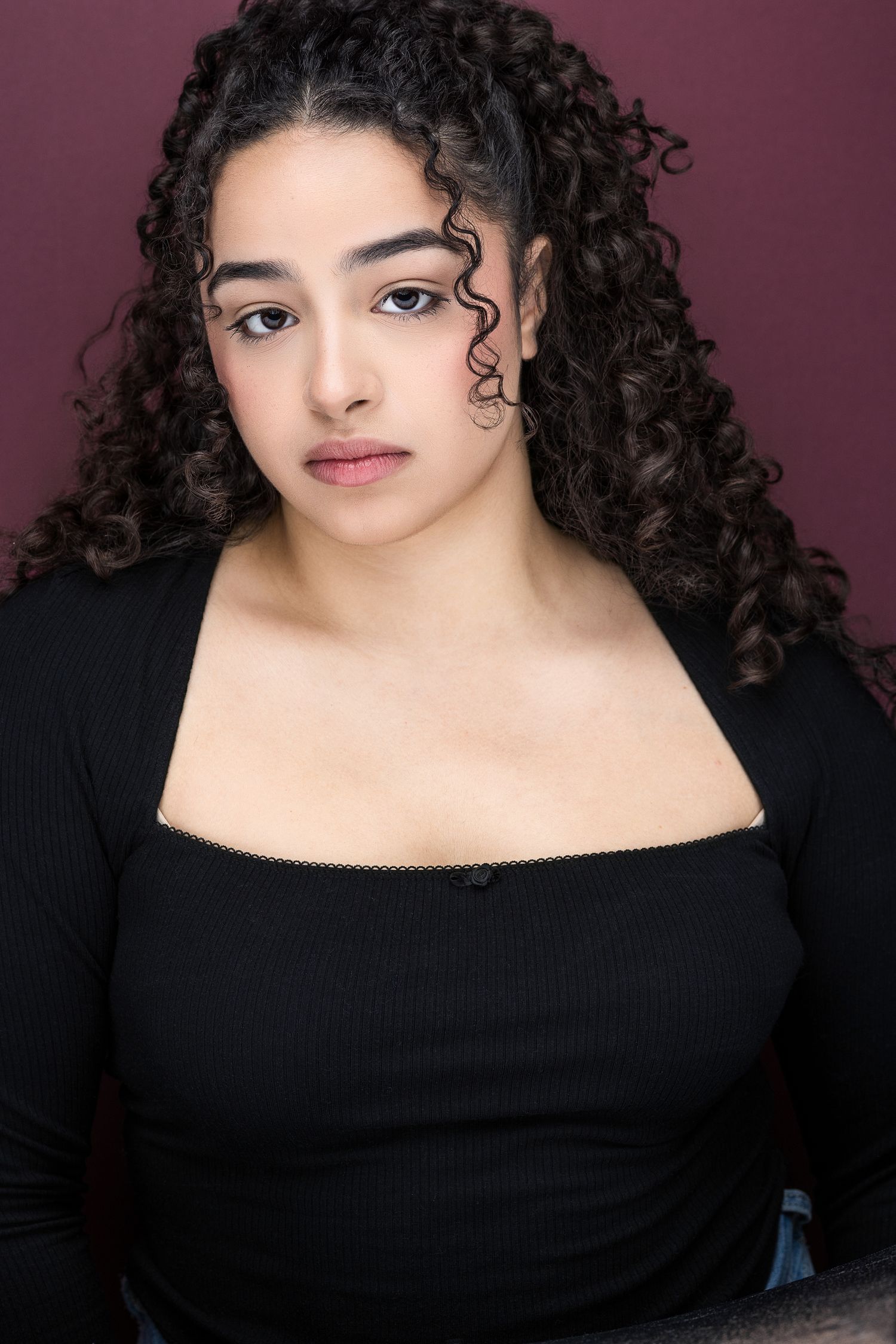 Young woman with curly black hair wearing black top against burgundy background.