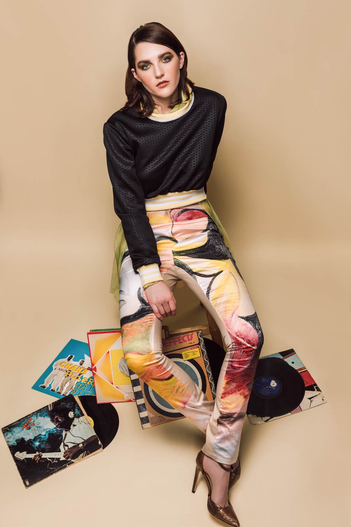 A stylish woman in colorful printed pants and black top poses with vintage vinyl records on a beige background.