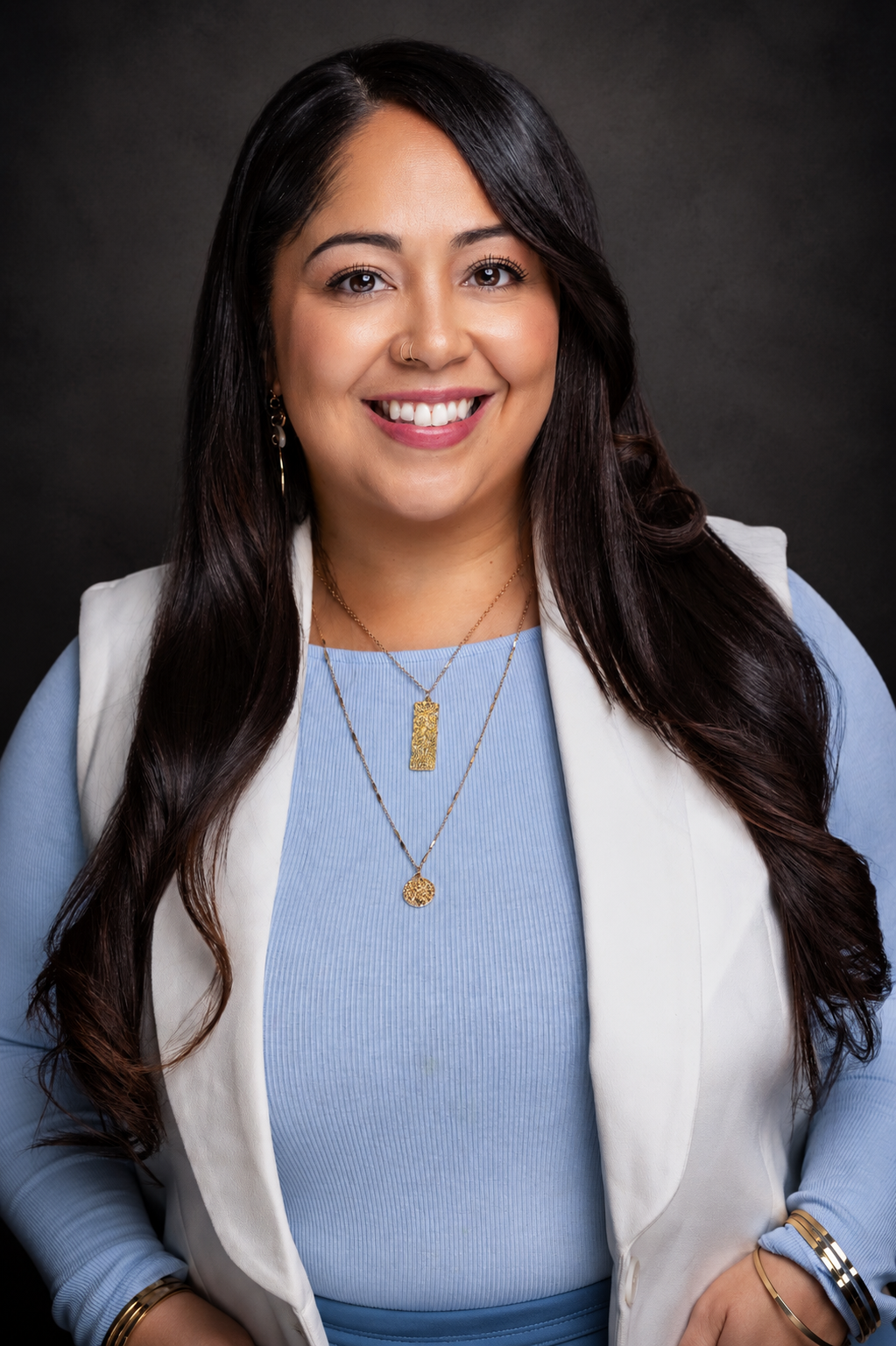 Professional headshot of a smiling woman with long dark hair wearing a blue top, white blazer, and gold necklaces.