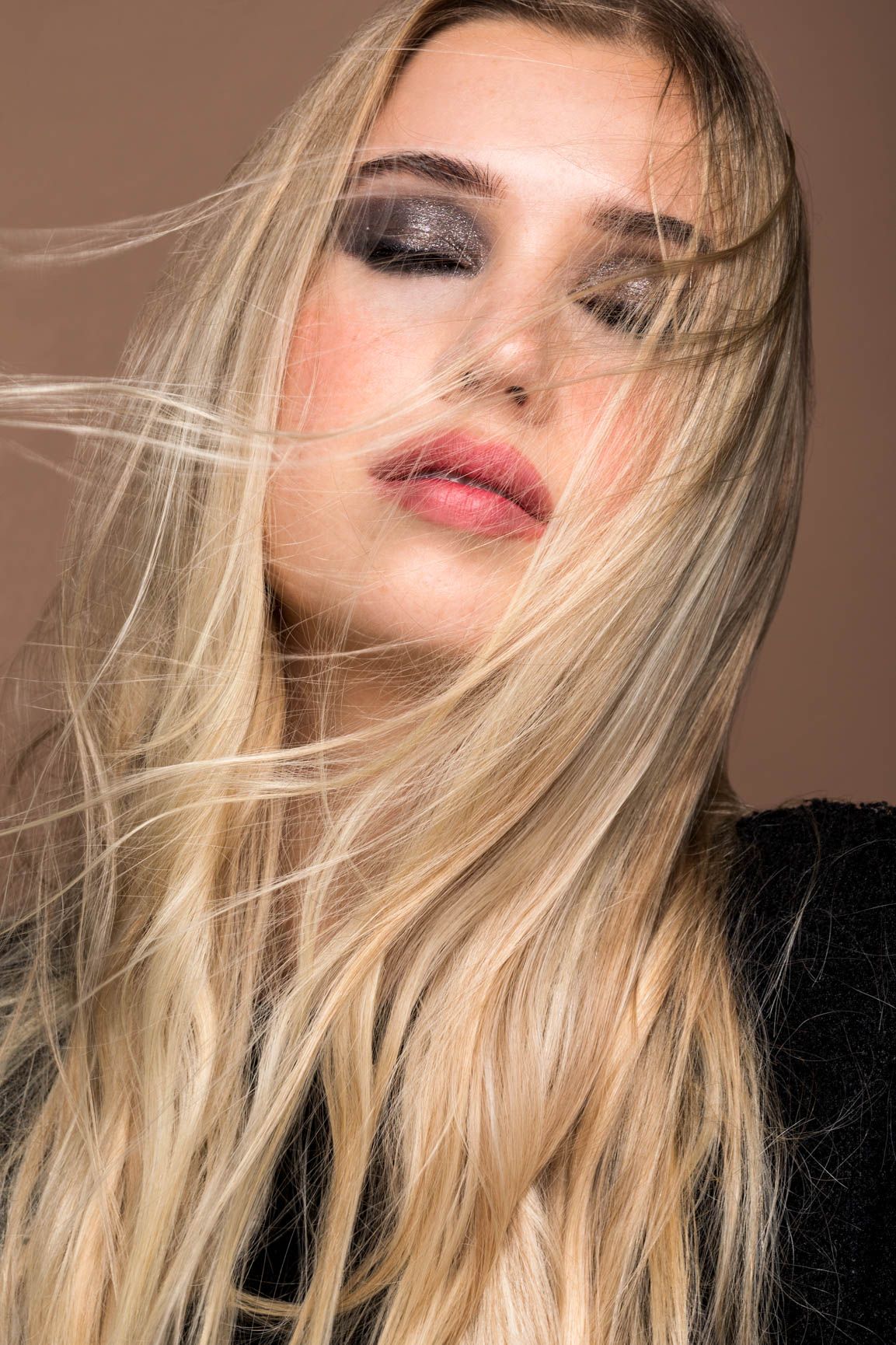 Blonde woman with smoky eye makeup and pink lips, hair blowing across face against brown background.