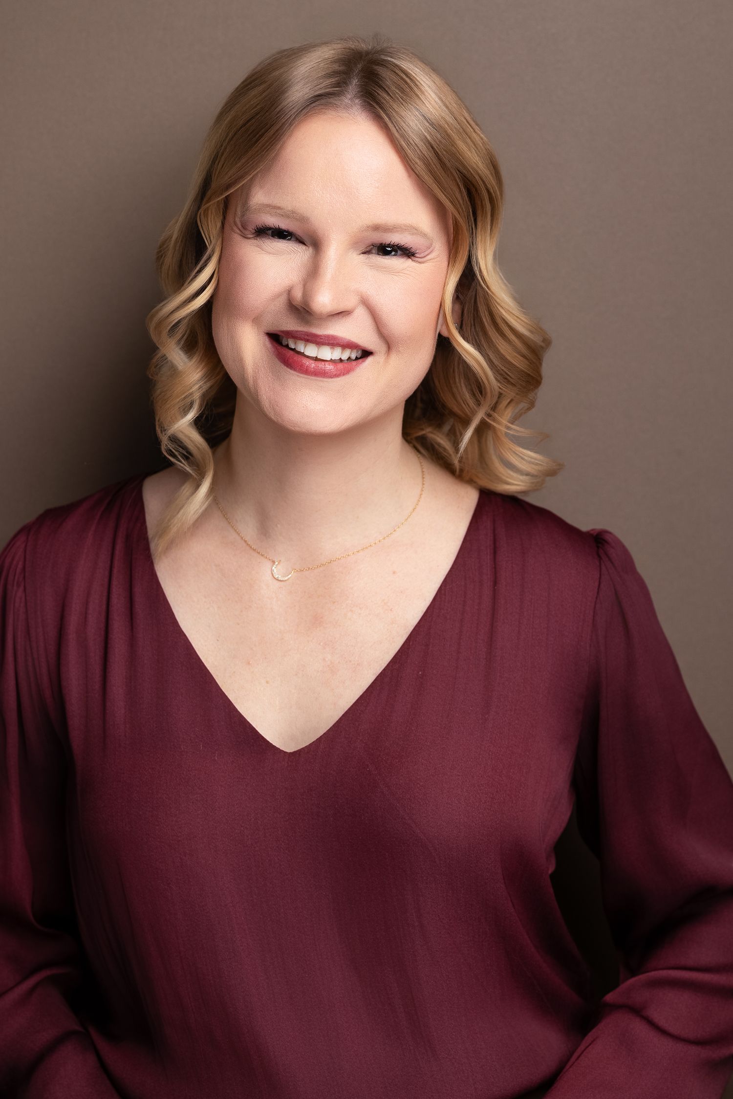 Smiling blonde woman in burgundy V-neck top with delicate necklace, professional studio headshot.