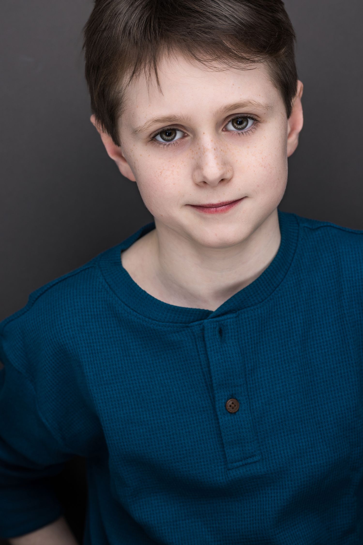 Young boy with short brown hair wearing a teal henley shirt poses for a professional headshot photo.