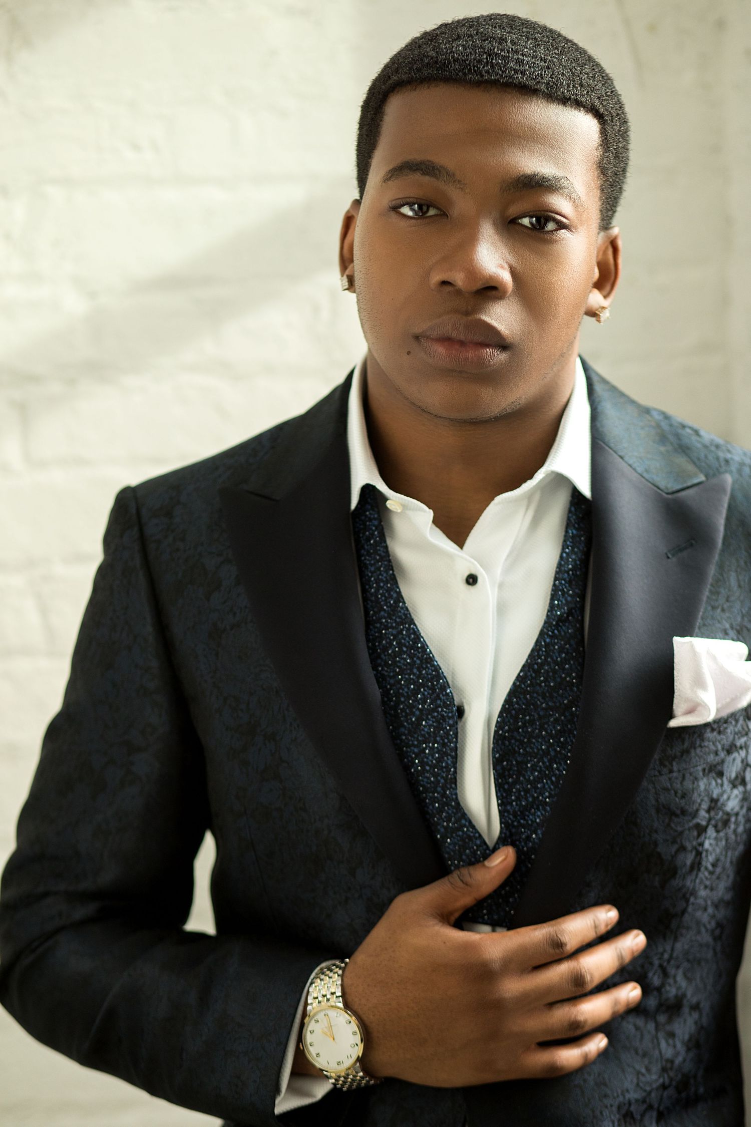Stylish young Black man in a navy patterned suit and gold watch poses confidently against a white brick wall.