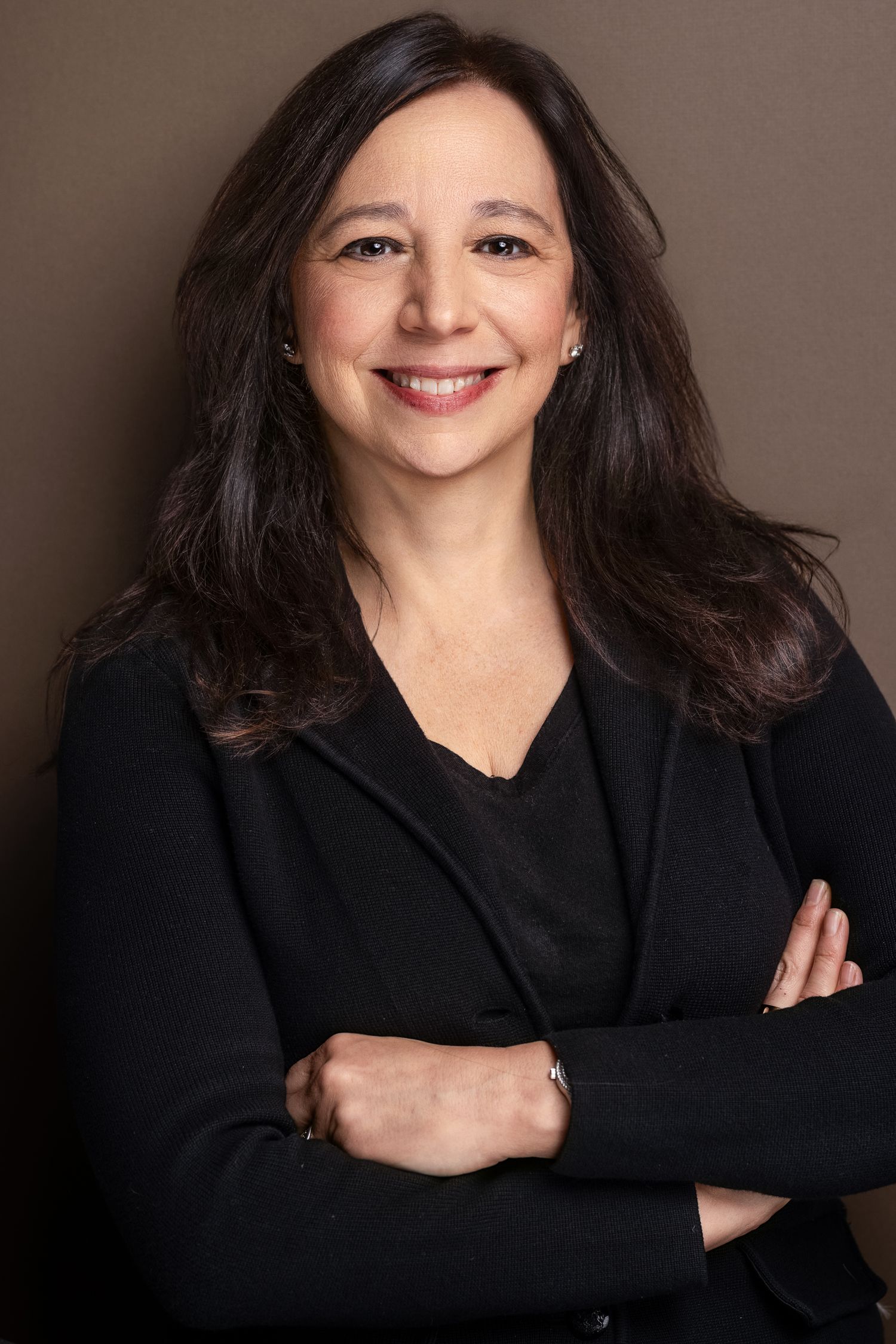 Professional headshot of a smiling woman with dark hair in a black blazer with arms crossed against brown backdrop.