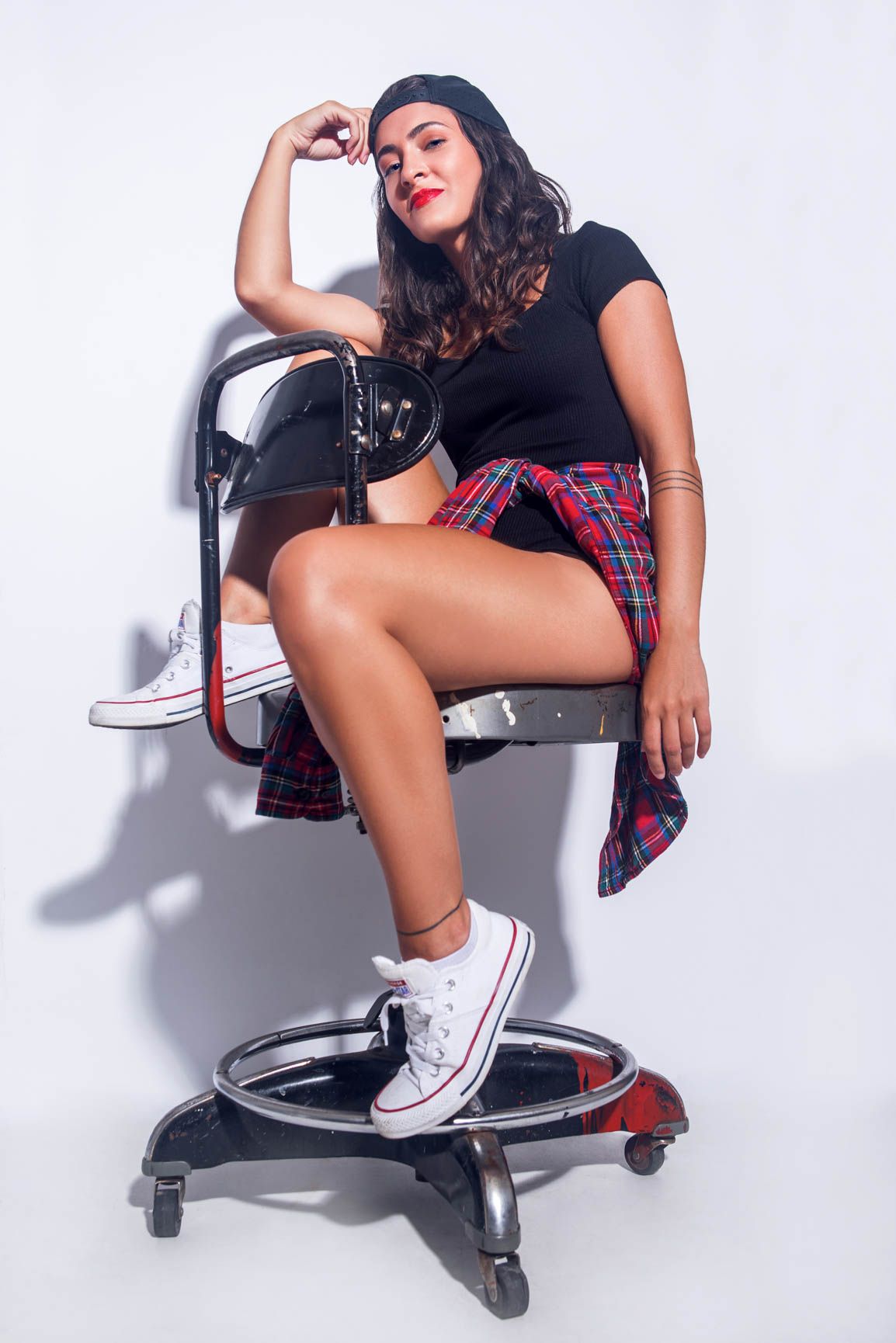 Young woman in plaid skirt and black top posing confidently on chair wearing white Converse sneakers.