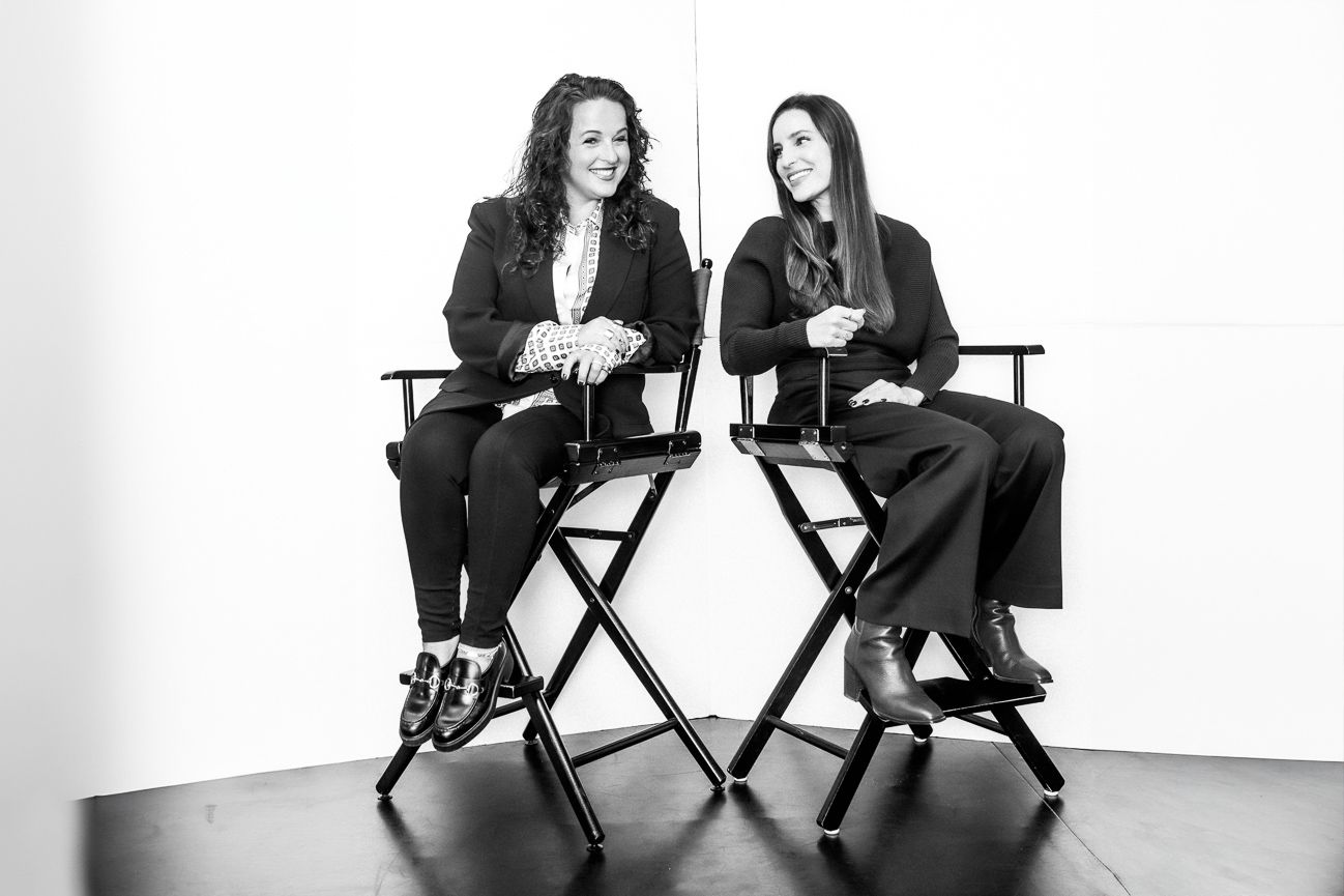 Two women in black blazers sitting in director chairs, smiling, black and white photo.