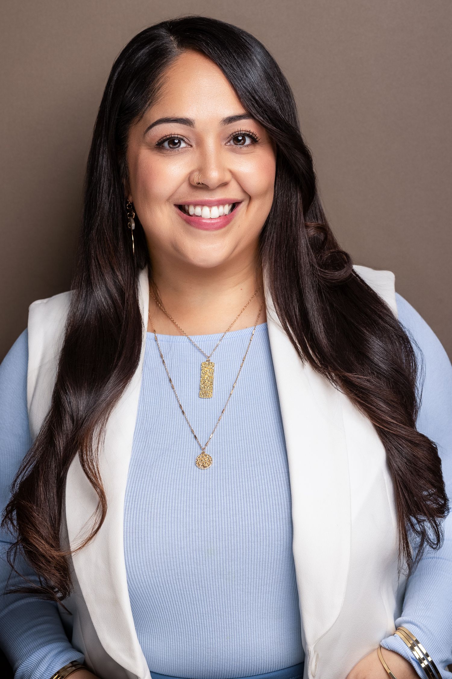 Smiling Latina woman with long dark hair in light blue top and white vest, professional business headshot.