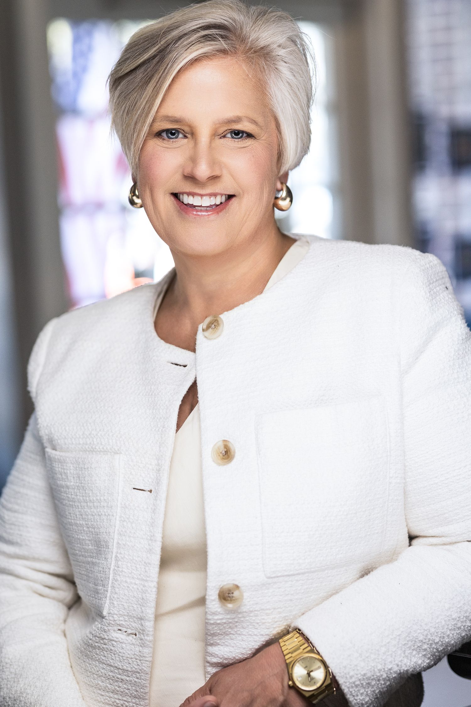 Professional headshot of a smiling woman with short gray hair wearing a white tweed blazer and gold jewelry.