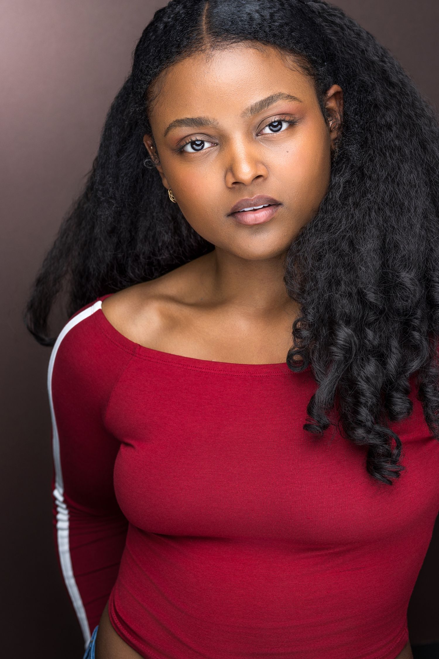 Young Black woman with long curly hair wearing a red off-shoulder top poses for a professional headshot.
