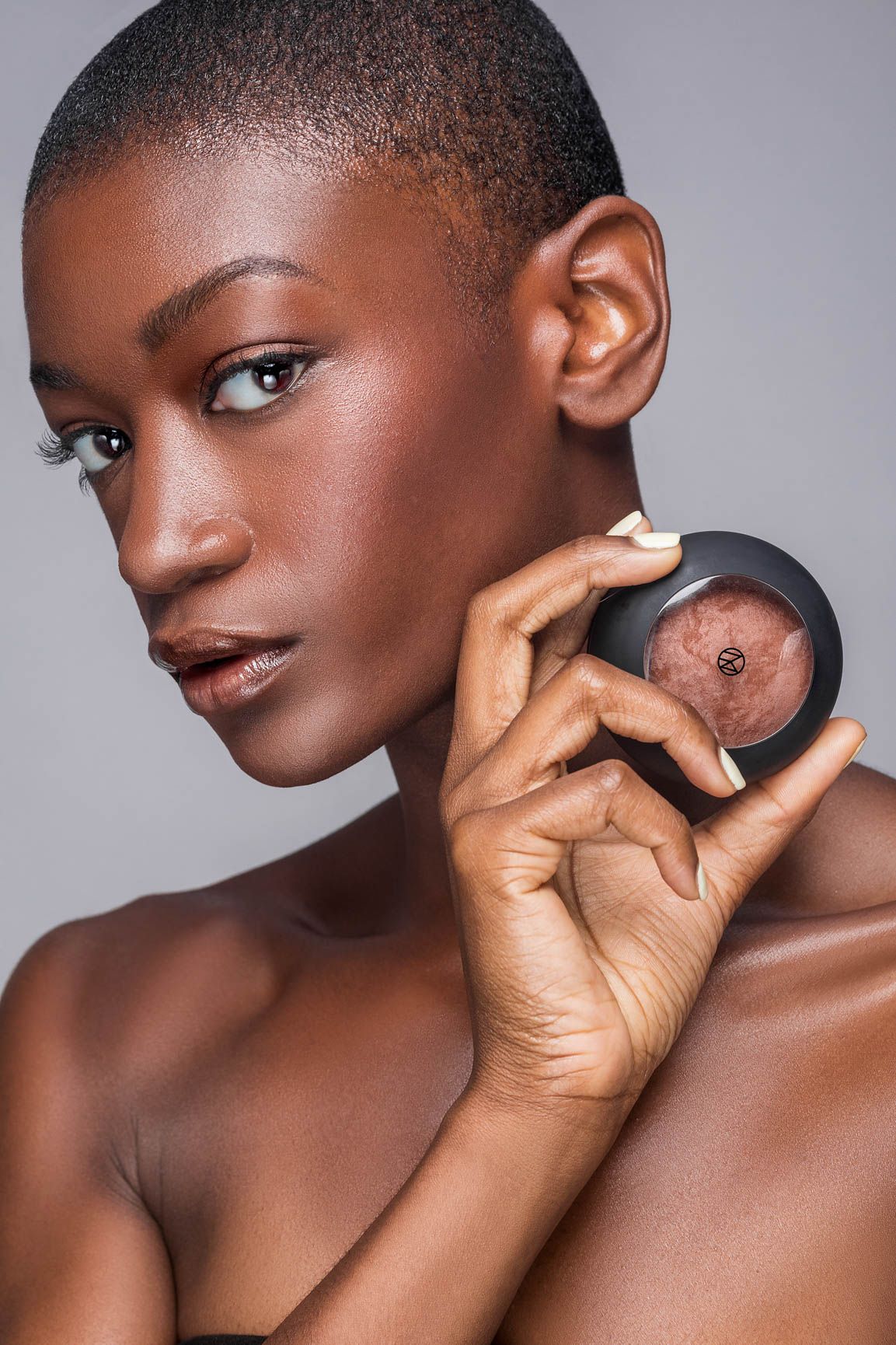 Elegant Black woman with shaved head holding makeup compact against gray background in beauty portrait.