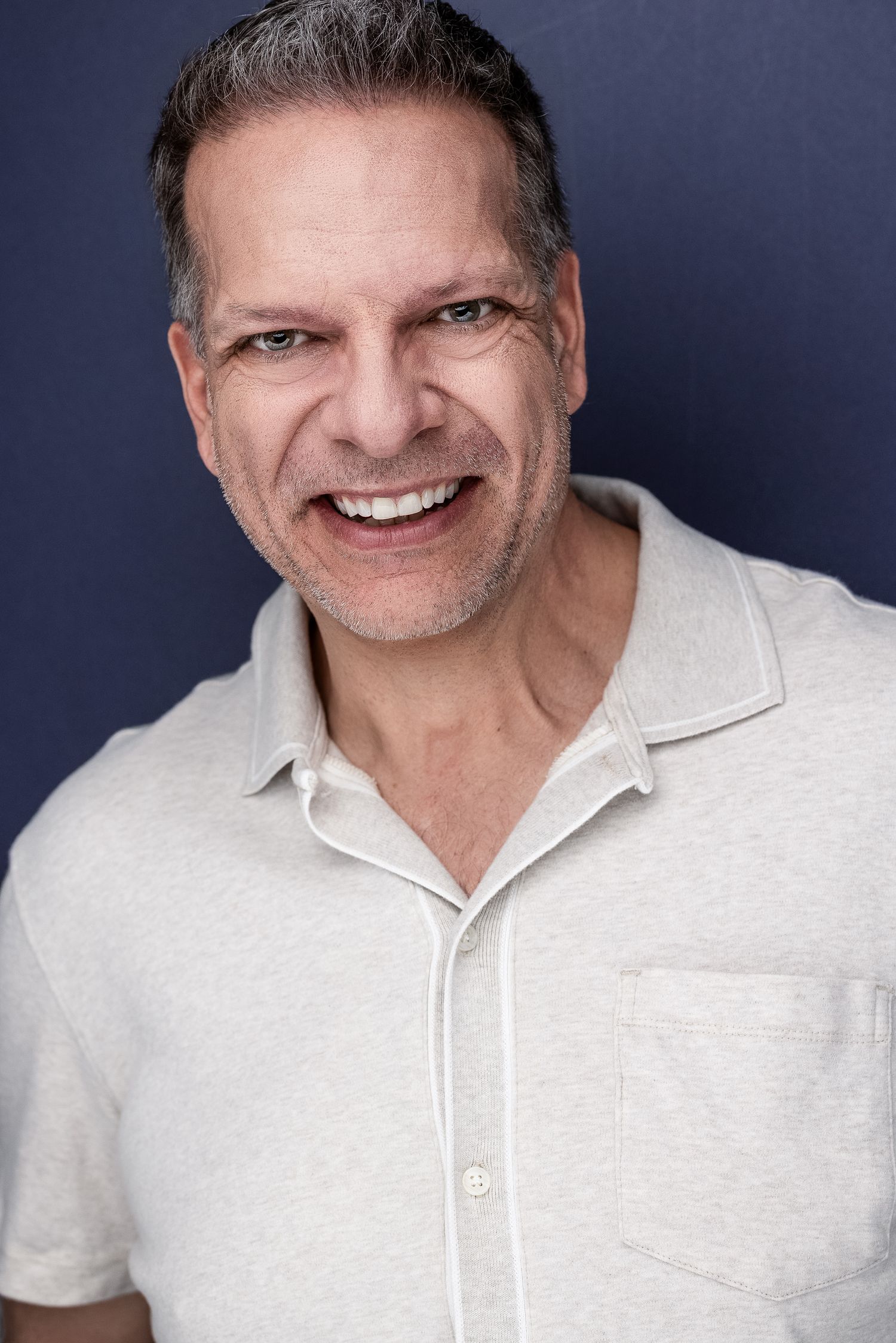 Middle-aged man with gray hair smiles broadly in white polo shirt against dark blue background.