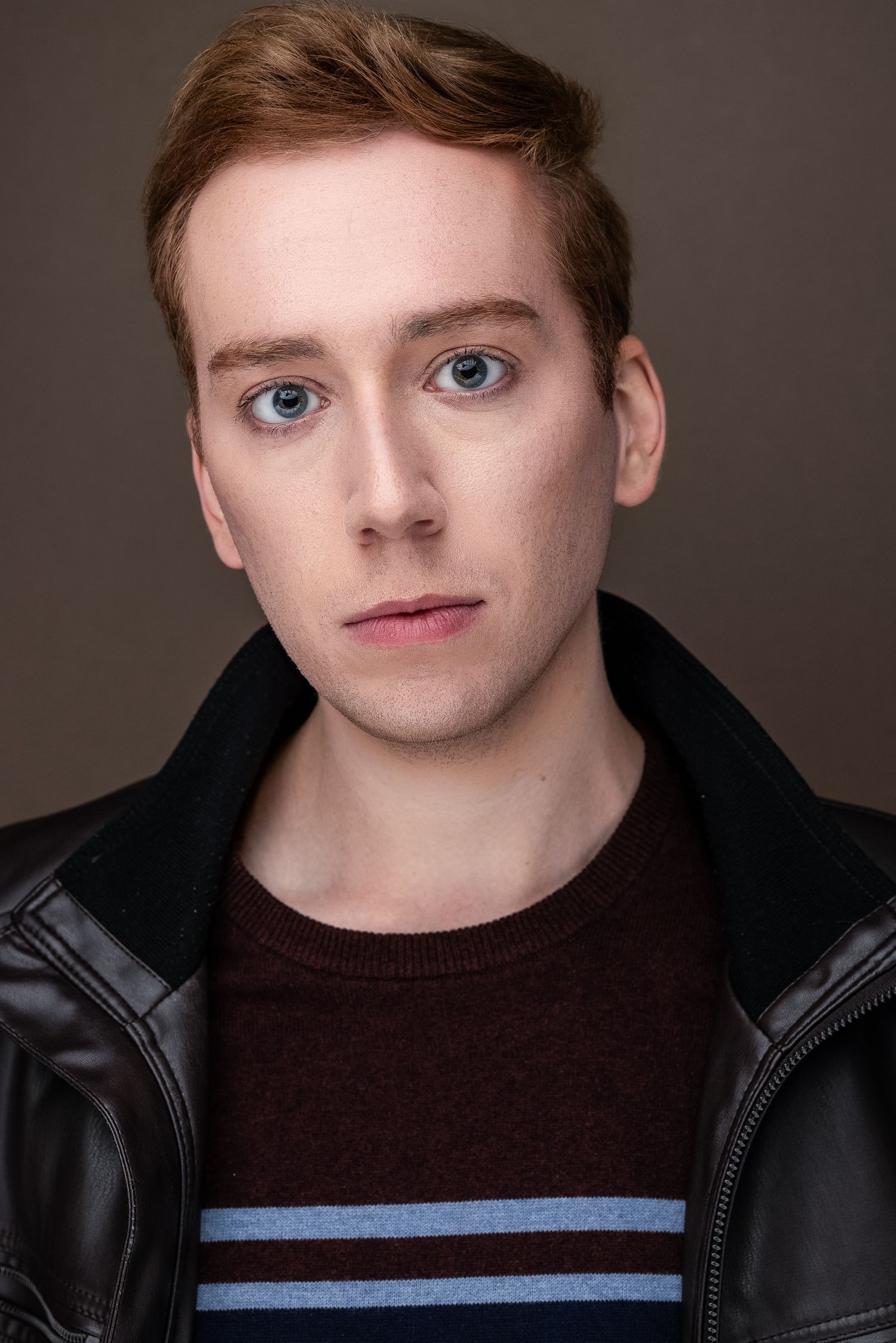 Young man with red hair and blue eyes wearing a leather jacket over a striped sweater in a professional headshot.