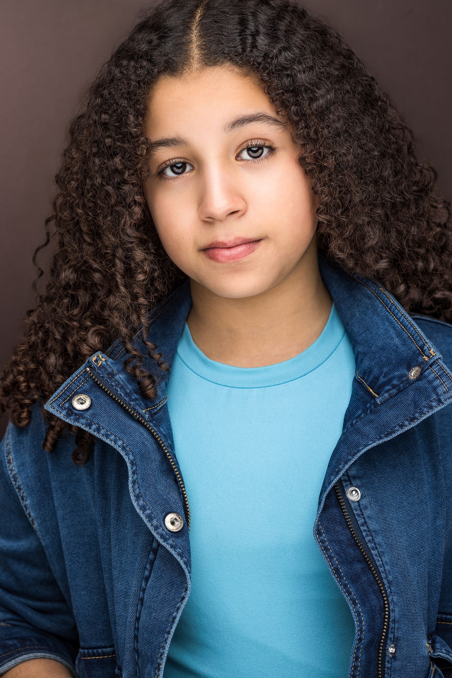 Young girl with curly hair wearing blue shirt and denim jacket against brown background.