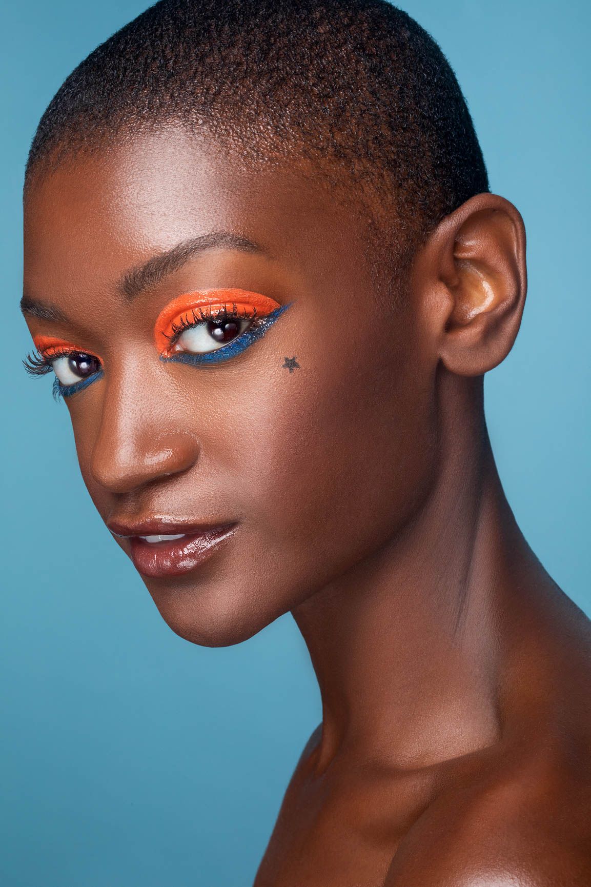 Bold beauty portrait of a woman with vibrant orange and blue eye makeup and a star tattoo on a blue background.