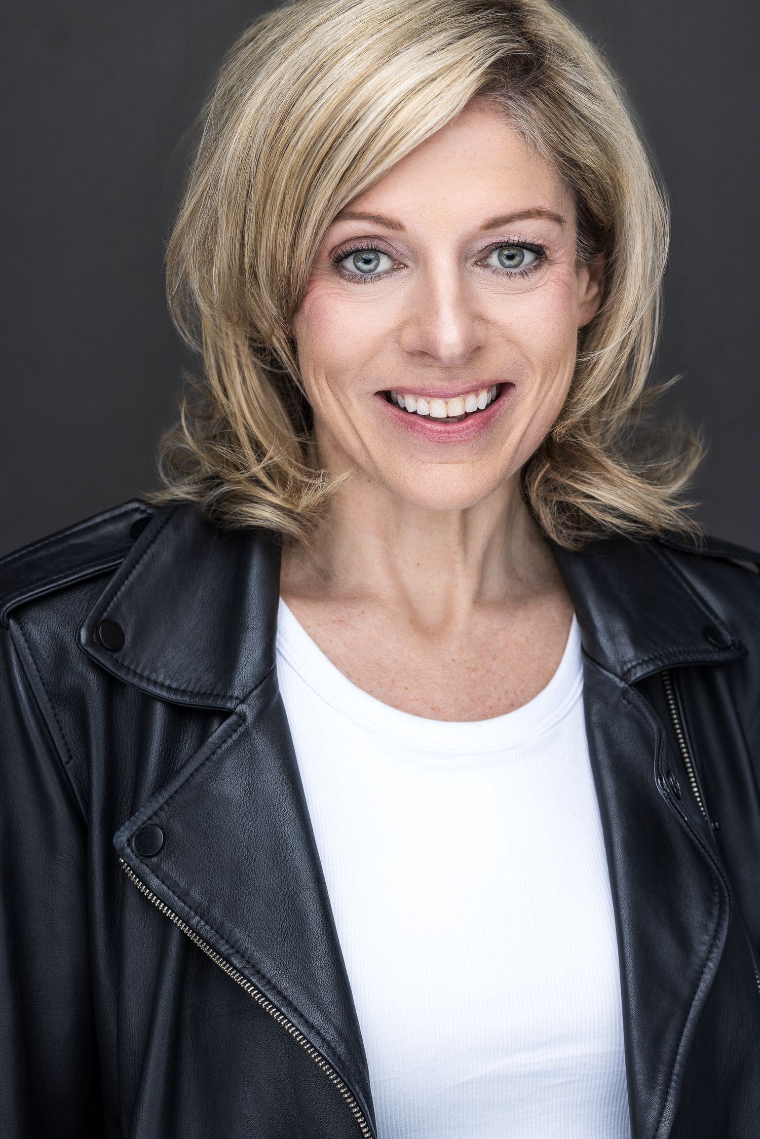 Blonde woman with blue eyes smiling in a black leather jacket and white shirt for a professional headshot.