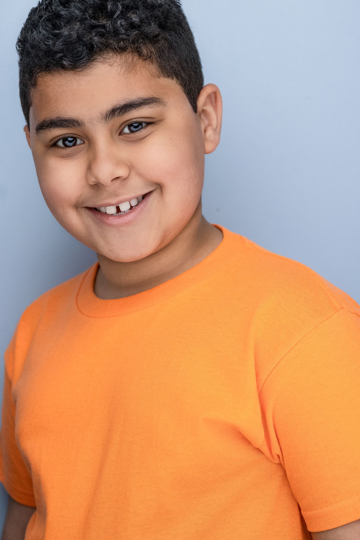 Smiling young boy with curly hair wearing an orange t-shirt against a light blue background.