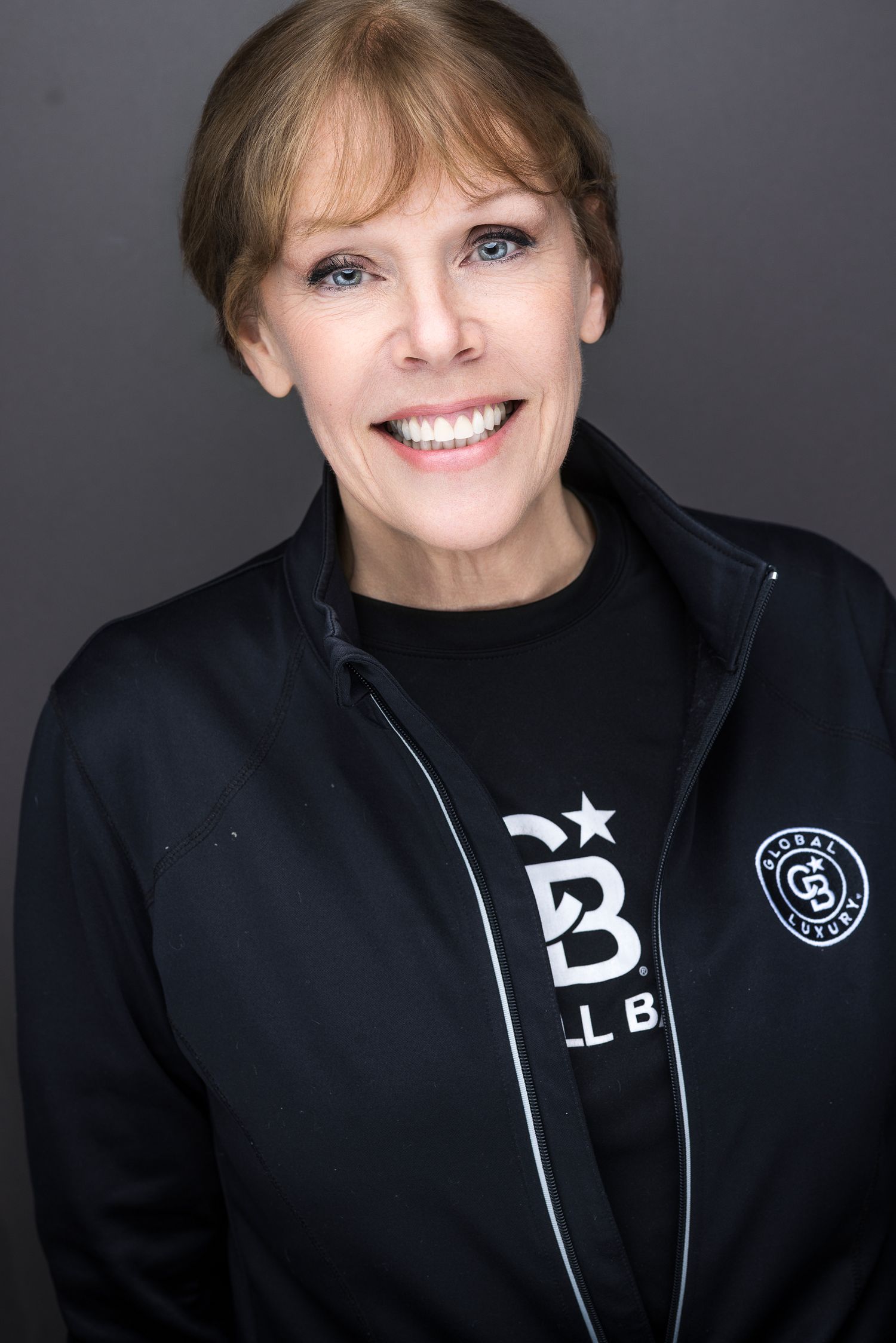 Smiling woman in black athletic jacket with logo, professional headshot on dark background.