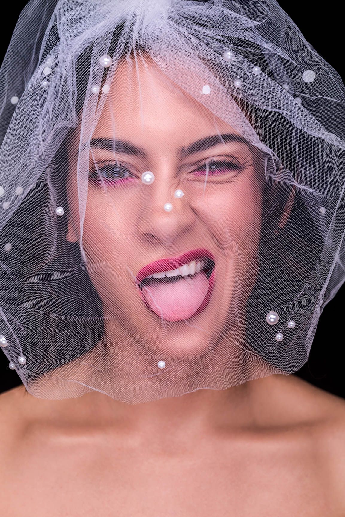 Playful bride sticking tongue out under pearl-embellished white veil with bold red lips on black background.