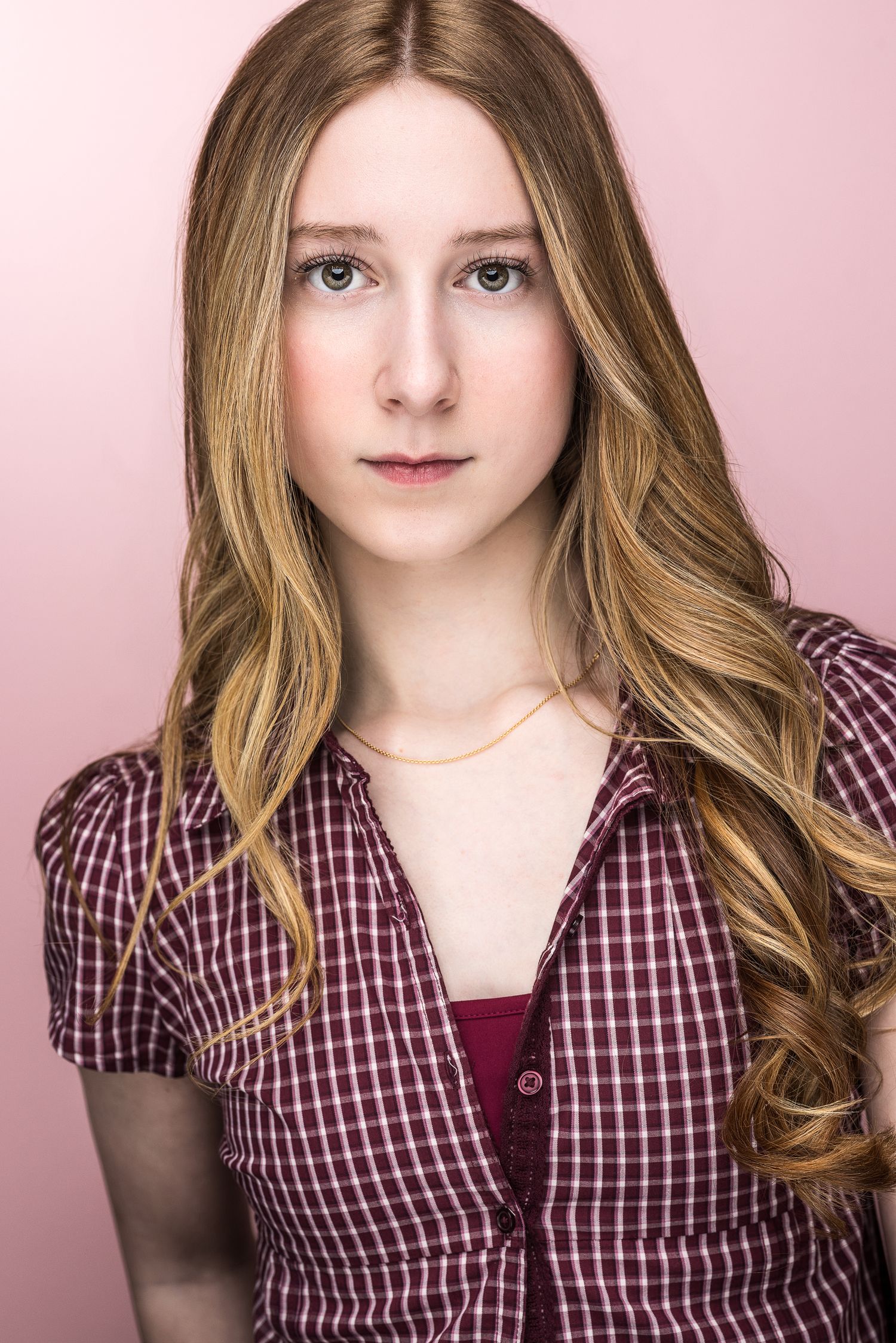 Young woman with long wavy blonde hair wearing a plaid shirt, posing against a pink background.