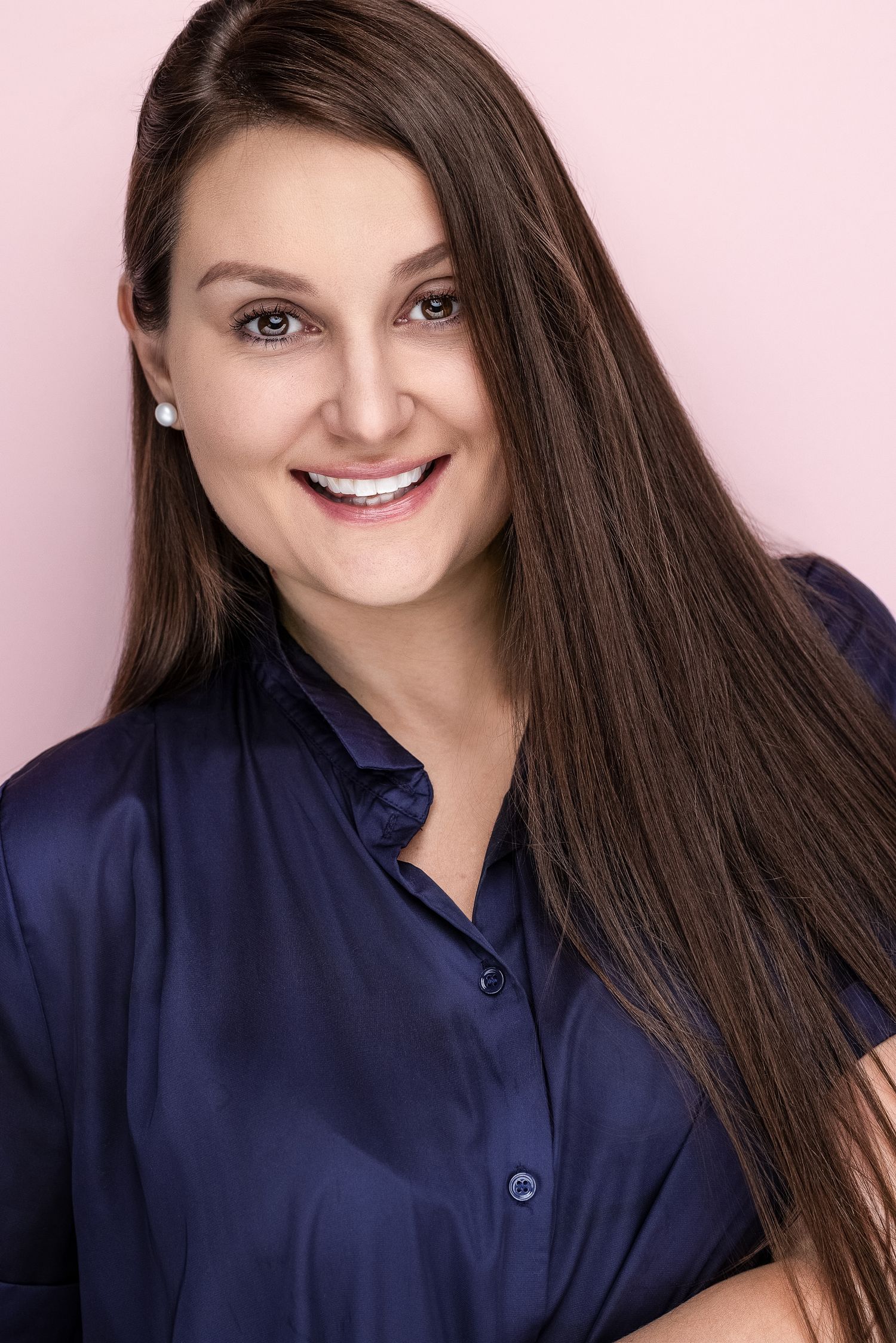 Smiling brunette woman in navy blue satin blouse with pearl earrings poses against pink background.