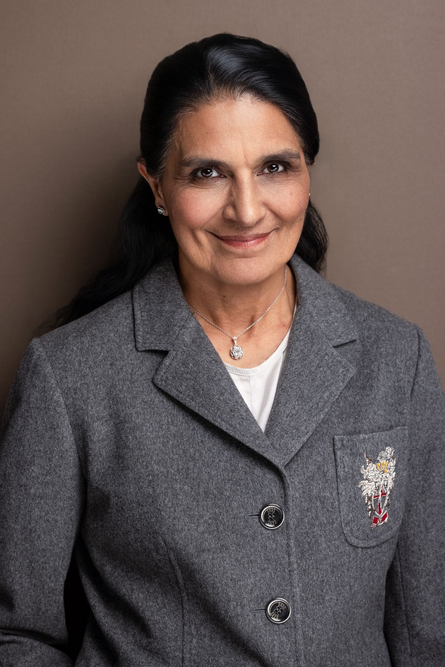 Professional headshot of a middle-aged woman in a gray blazer with floral brooch and necklace, smiling confidently.