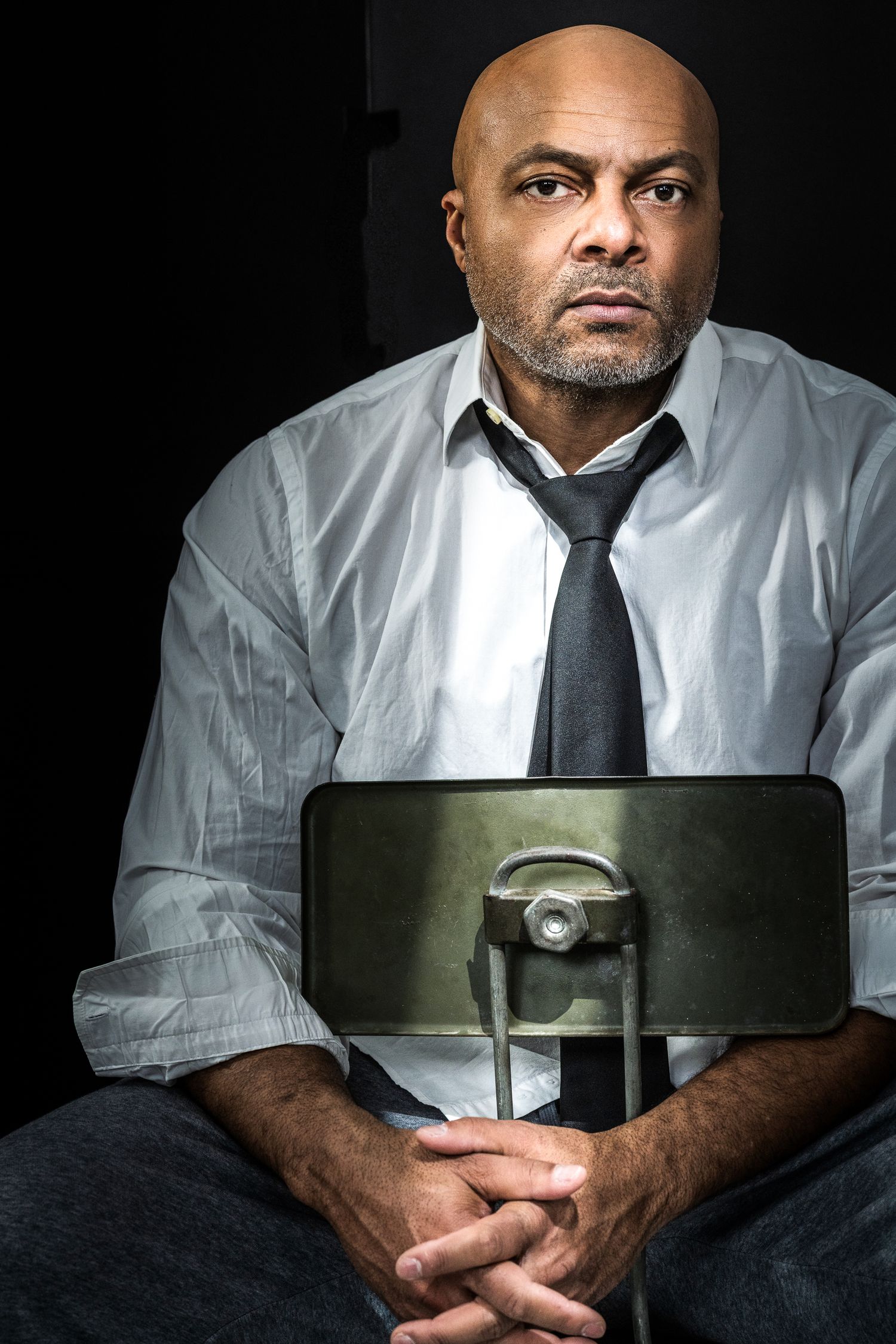 Bald Black man in white shirt and tie sitting with hands clasped in dramatic lighting.