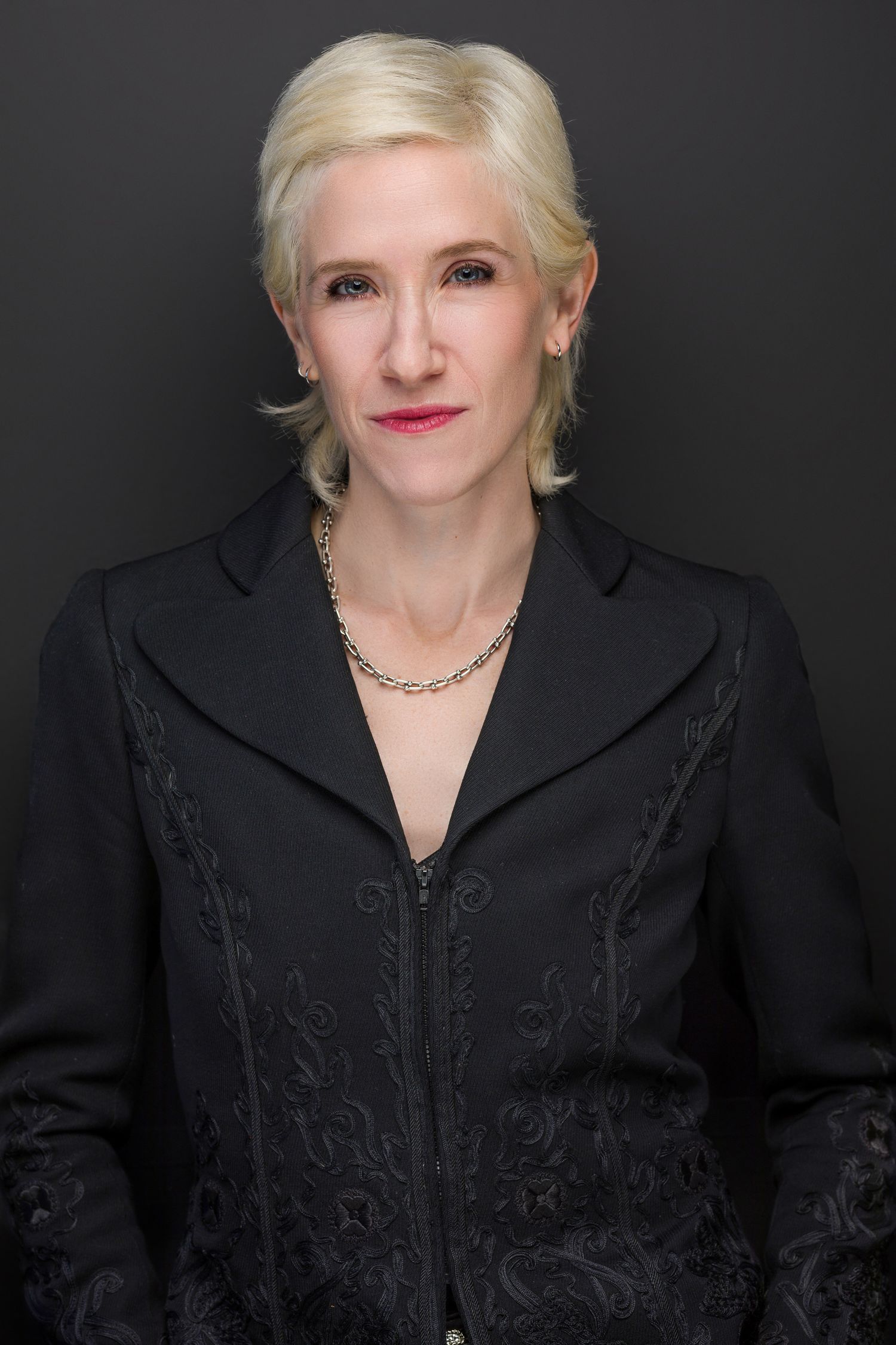 Professional woman with blonde hair wearing a black embroidered jacket and gold necklace poses against dark background.