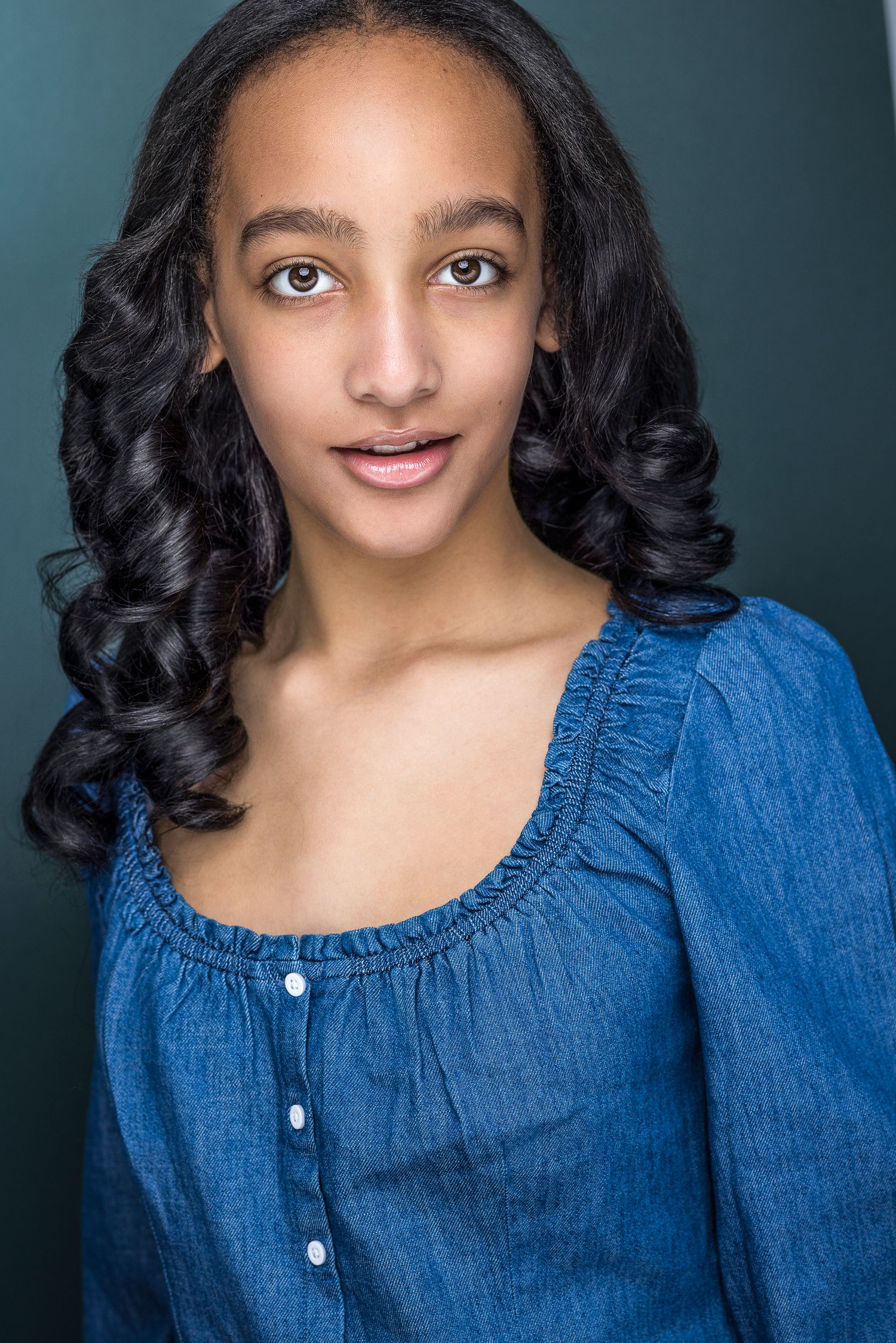 Young biracial teen girl with curly black hair in blue denim top poses for professional acting headshot.