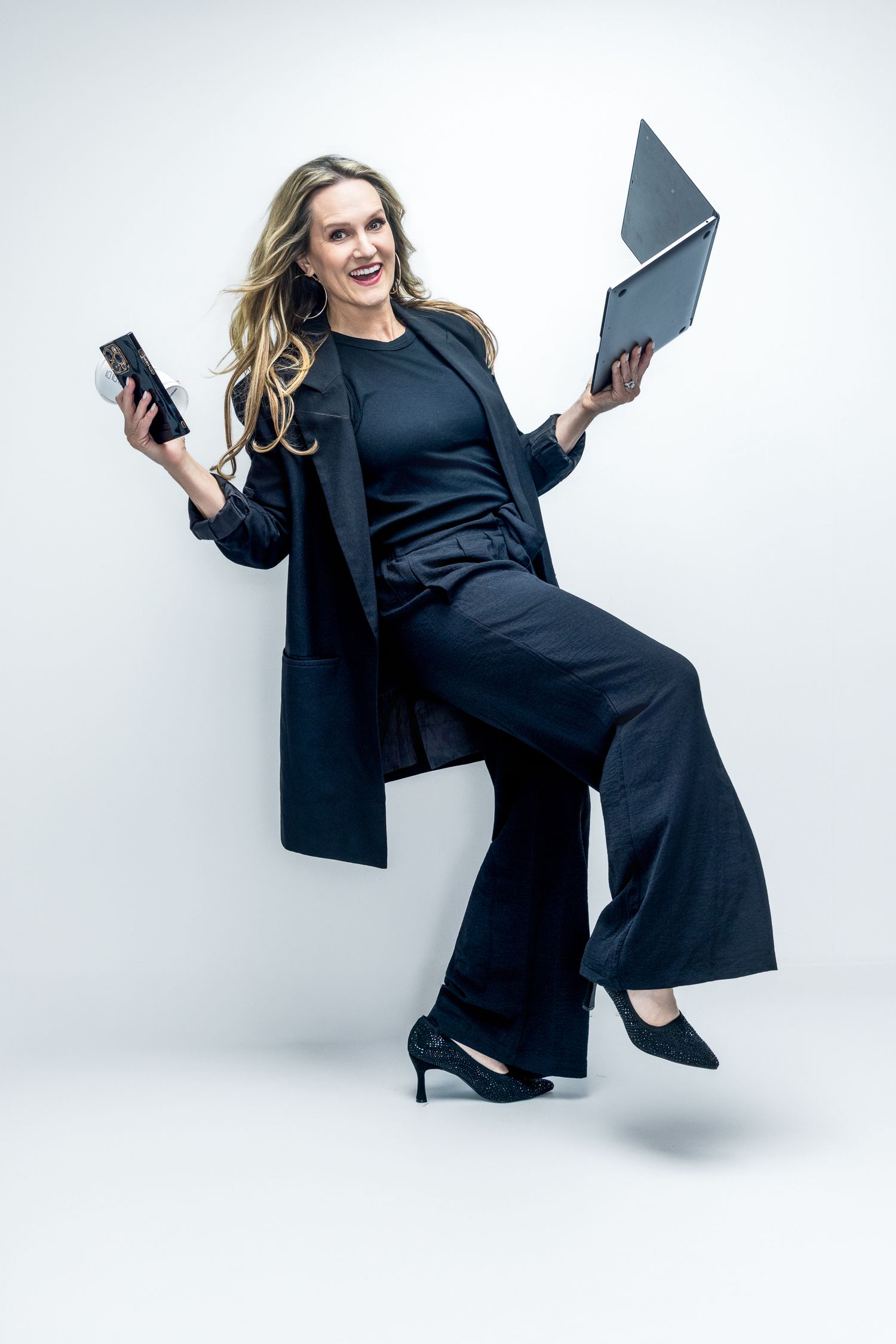 Smiling businesswoman in black suit holding laptop and phone against white background.