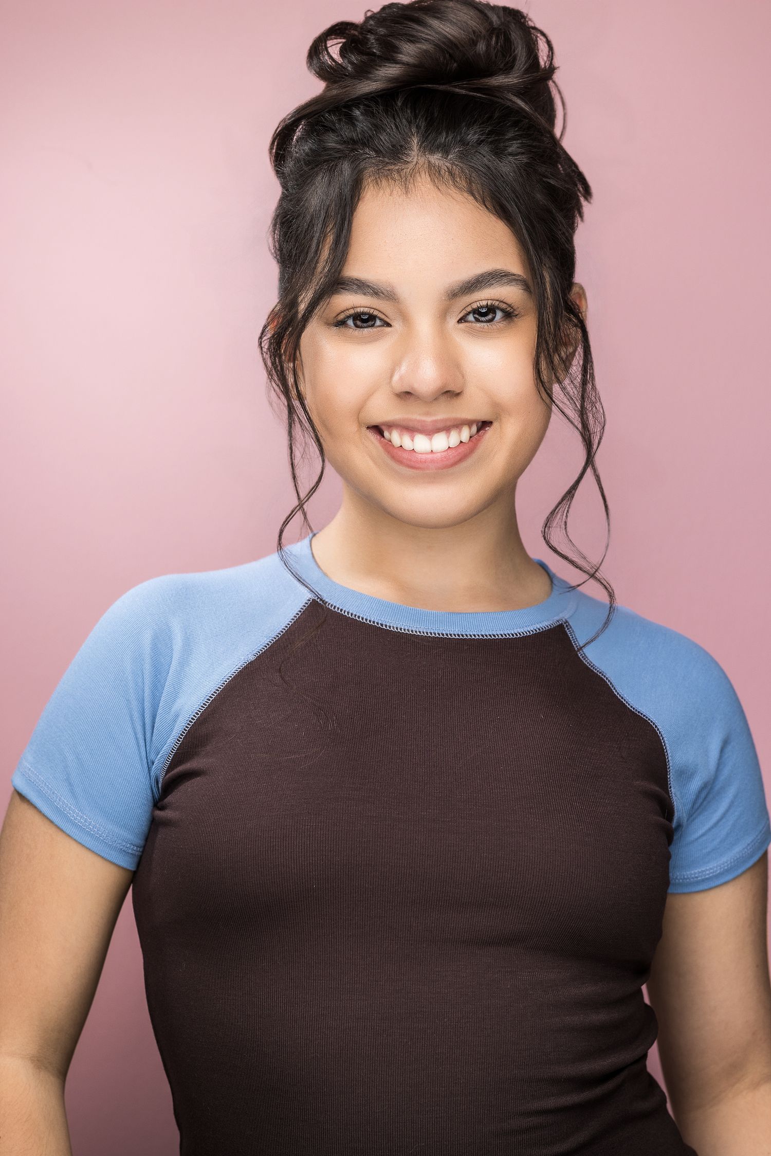 Smiling young woman with dark hair in a bun wearing a brown and blue raglan shirt against a pink background.