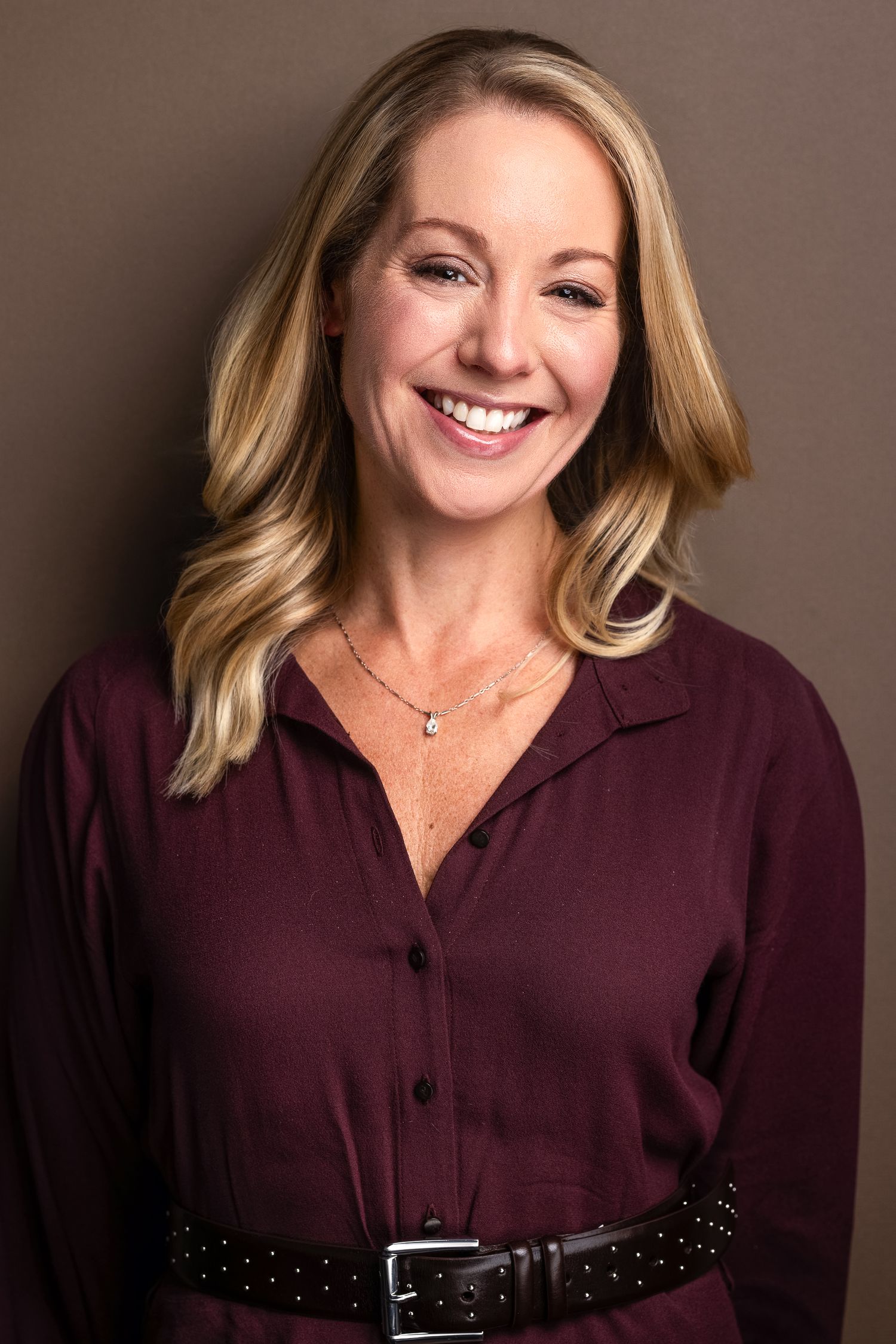 Smiling blonde woman in burgundy blouse with black belt poses for professional headshot photo.