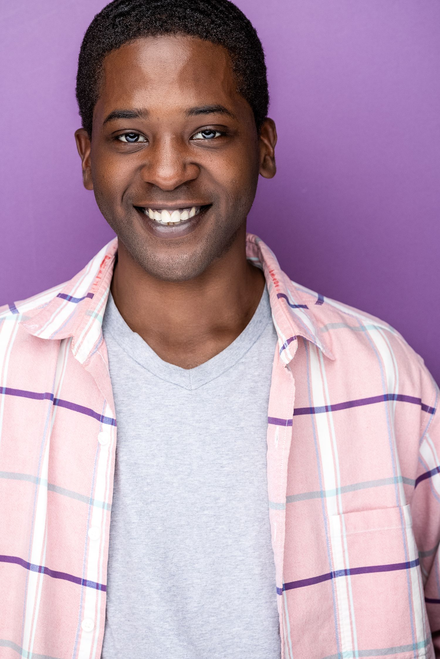Smiling young Black man wearing a pink plaid shirt over gray t-shirt, posing against a purple background.