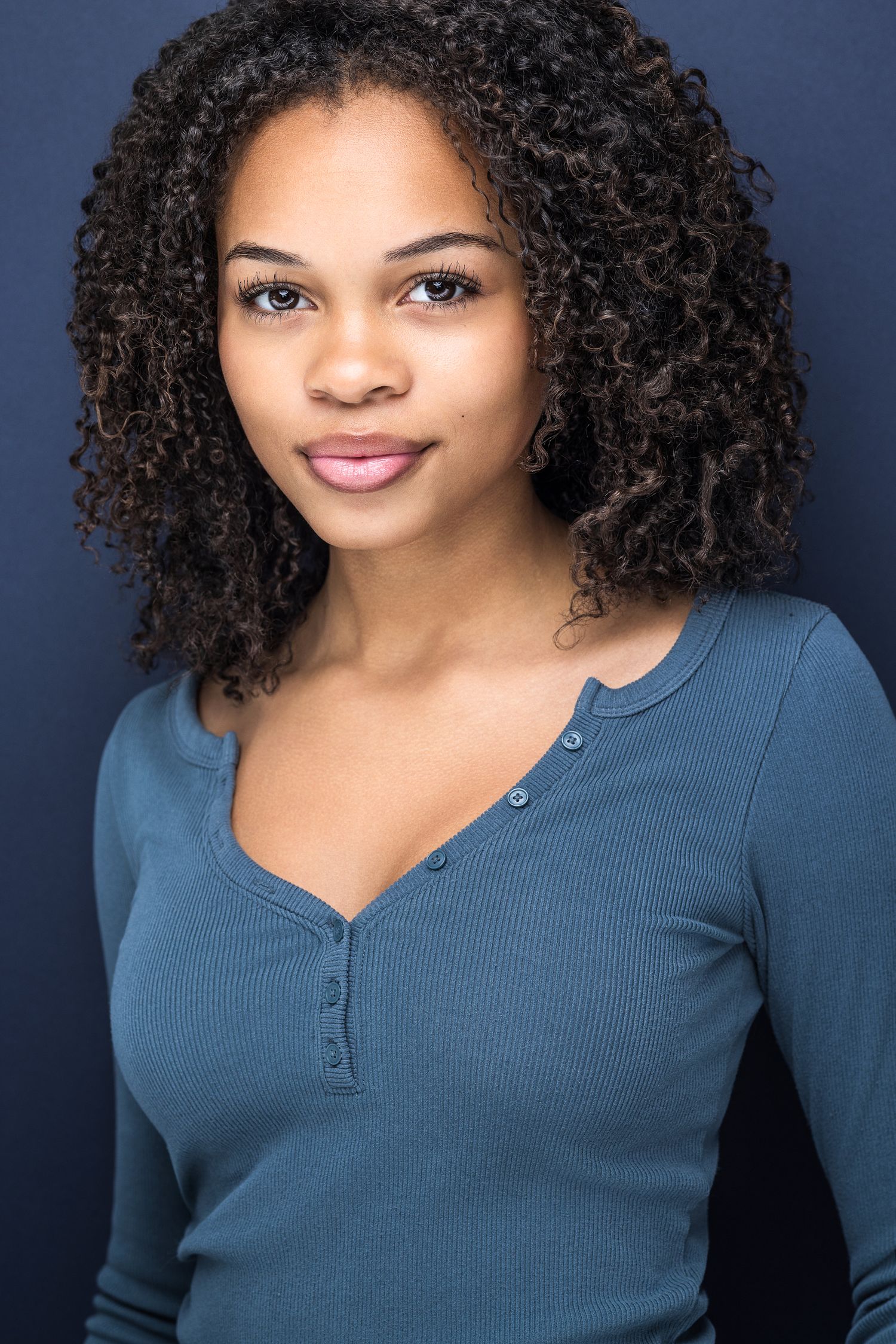 Young Black woman with natural curly hair wearing teal henley top smiles for professional acting headshot.
