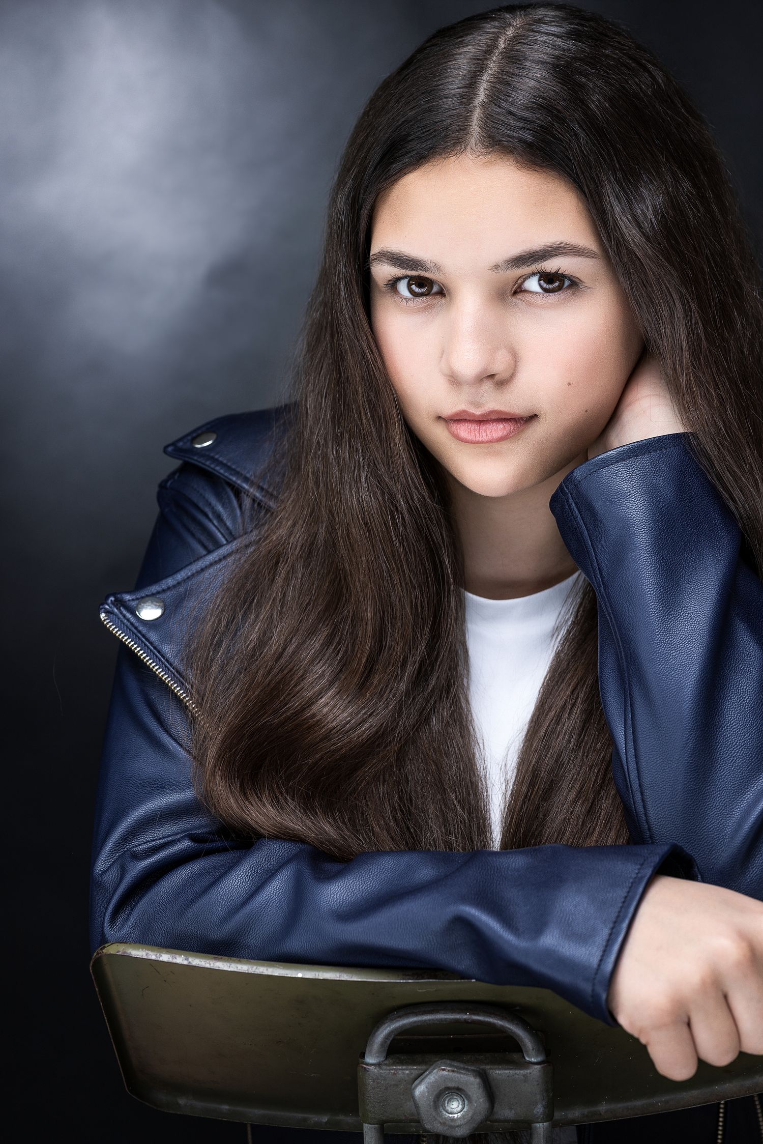 Young girl with long dark hair wearing a blue leather jacket, posing confidently against a dark studio background.