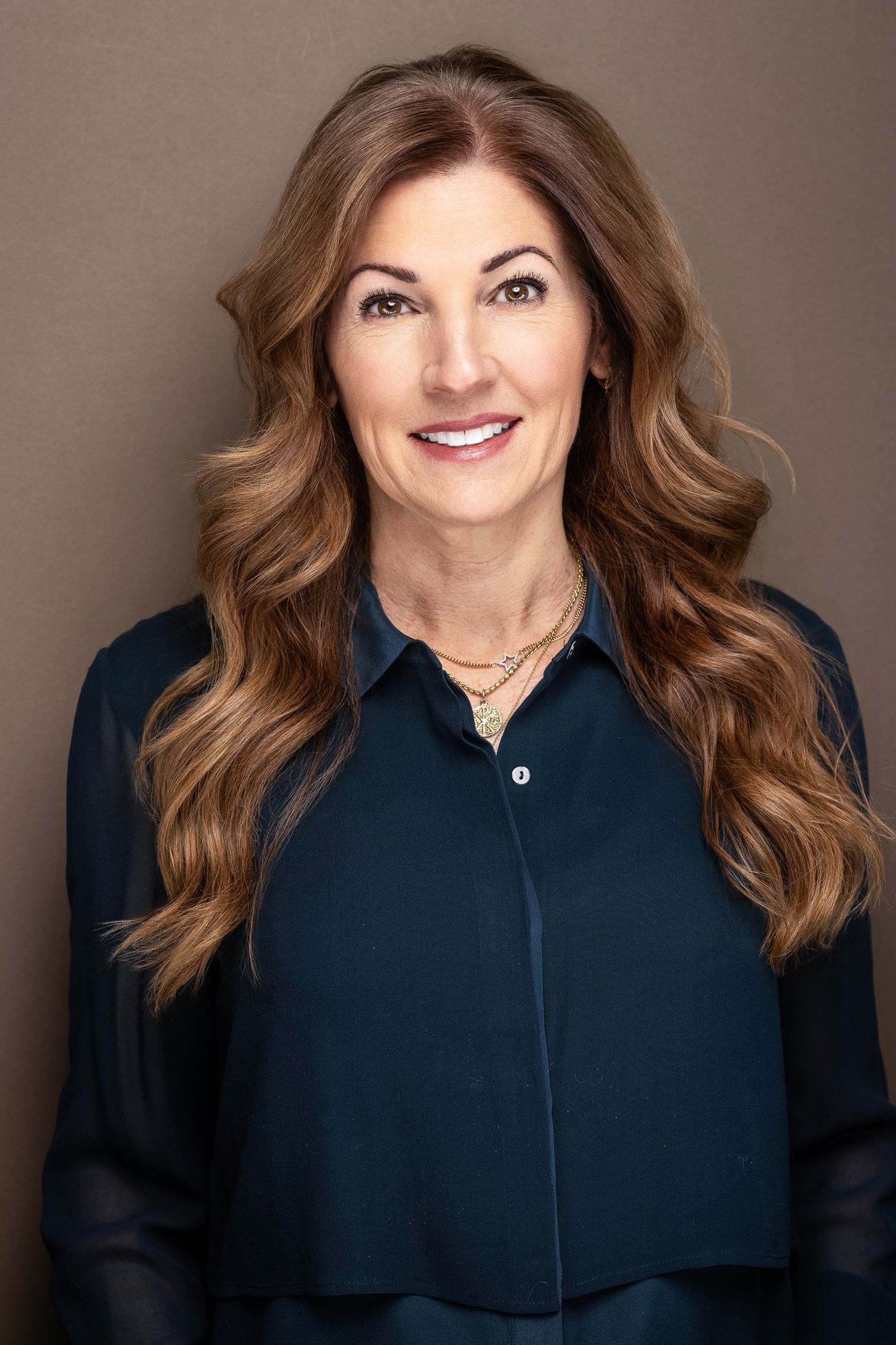 Woman with long wavy brown hair in navy blouse and gold necklace, professional headshot.