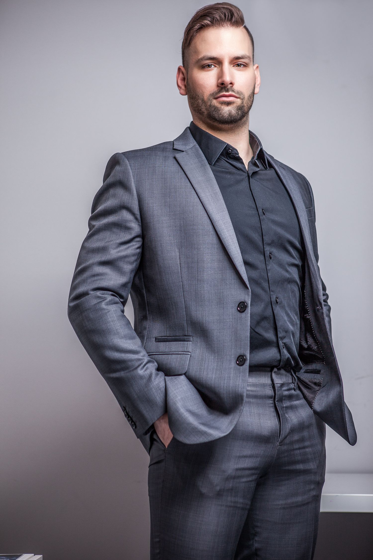 Confident businessman in gray suit and black shirt standing with hands in pockets against neutral background.