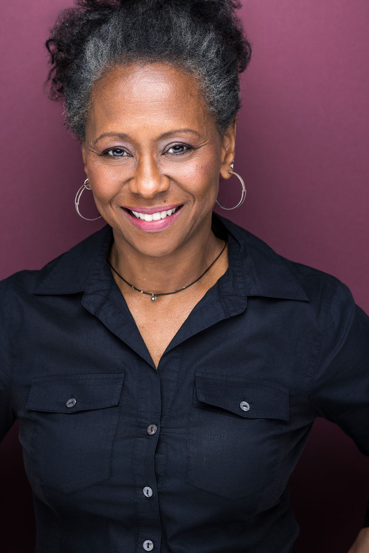 Confident older Black woman with gray hair wearing a black shirt and hoop earrings against a dark red background.