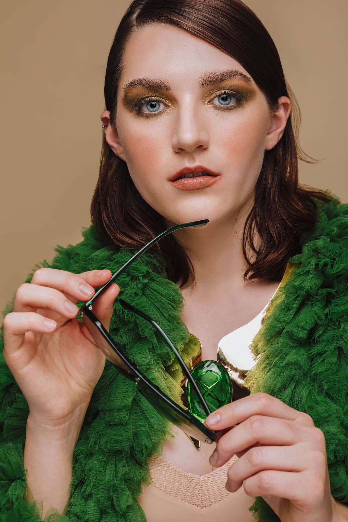 Young woman in green fluffy jacket holding sunglasses, wearing bold eye makeup, posing against beige background.