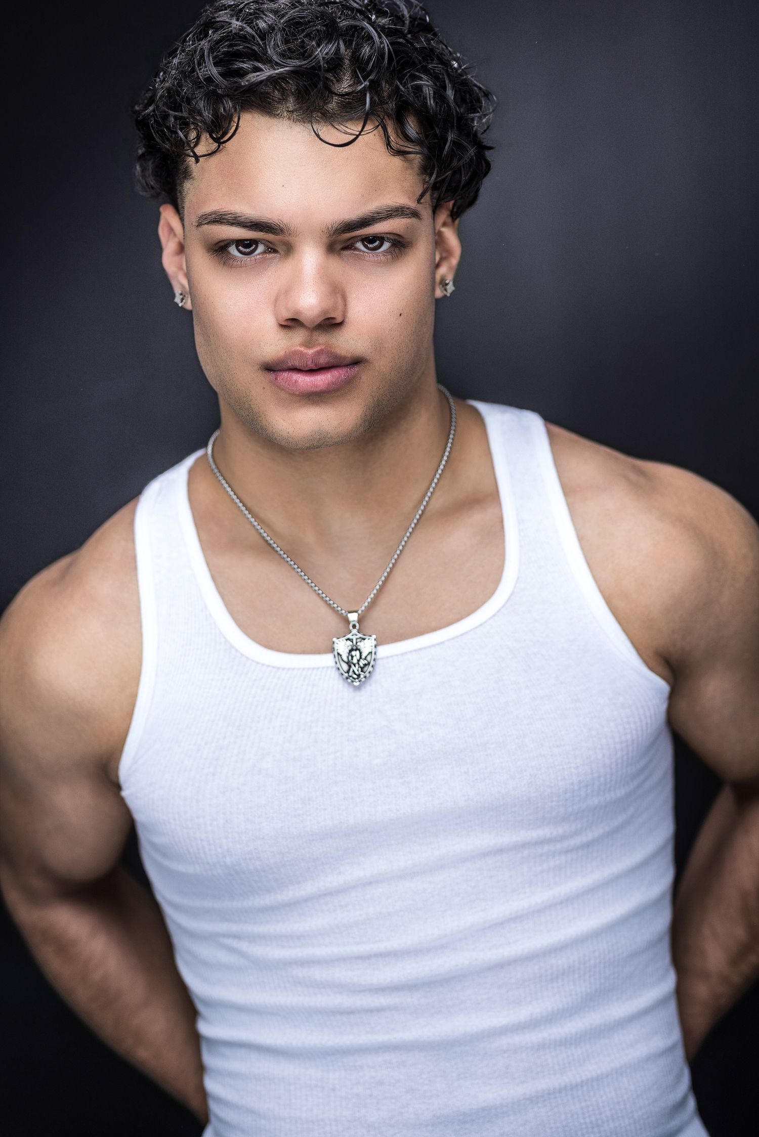 Muscular young man in white tank top with silver necklace poses for a professional fitness headshot.