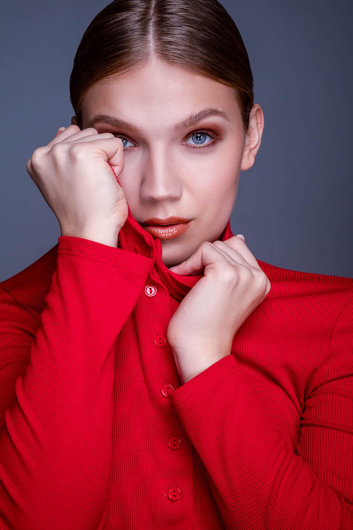 Young woman in red ribbed turtleneck shirt holding collar with both hands against gray background.