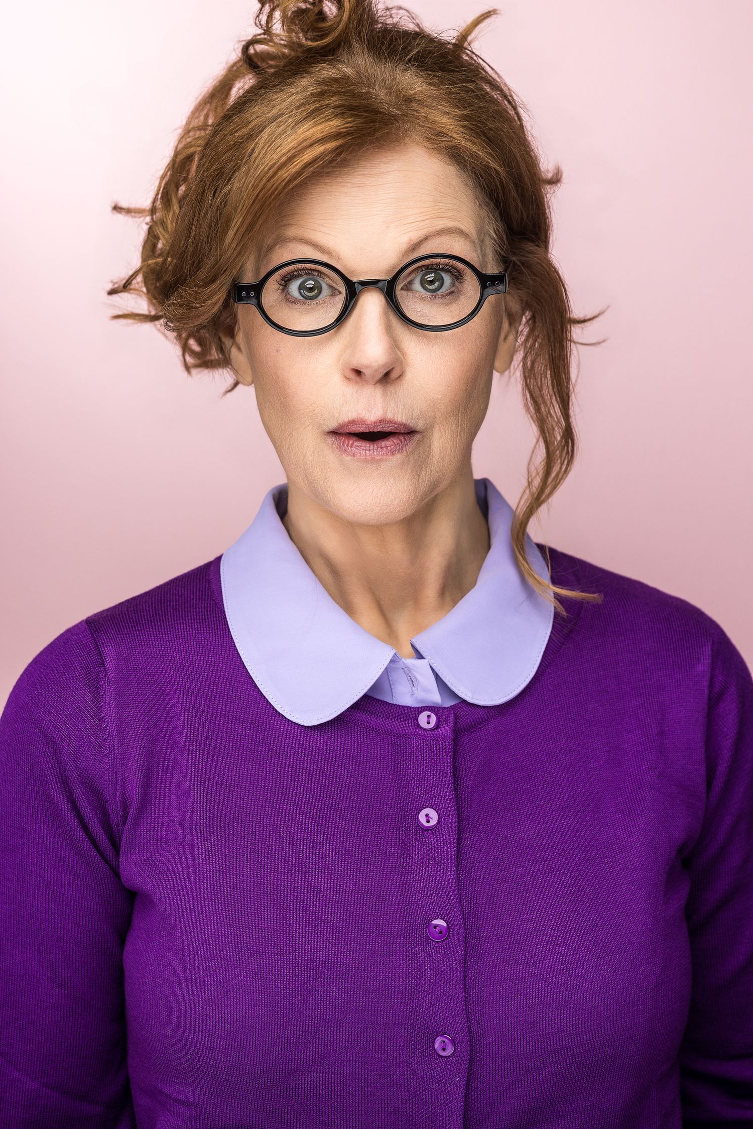 Surprised woman with red hair and round glasses wearing a purple cardigan against a pink background.