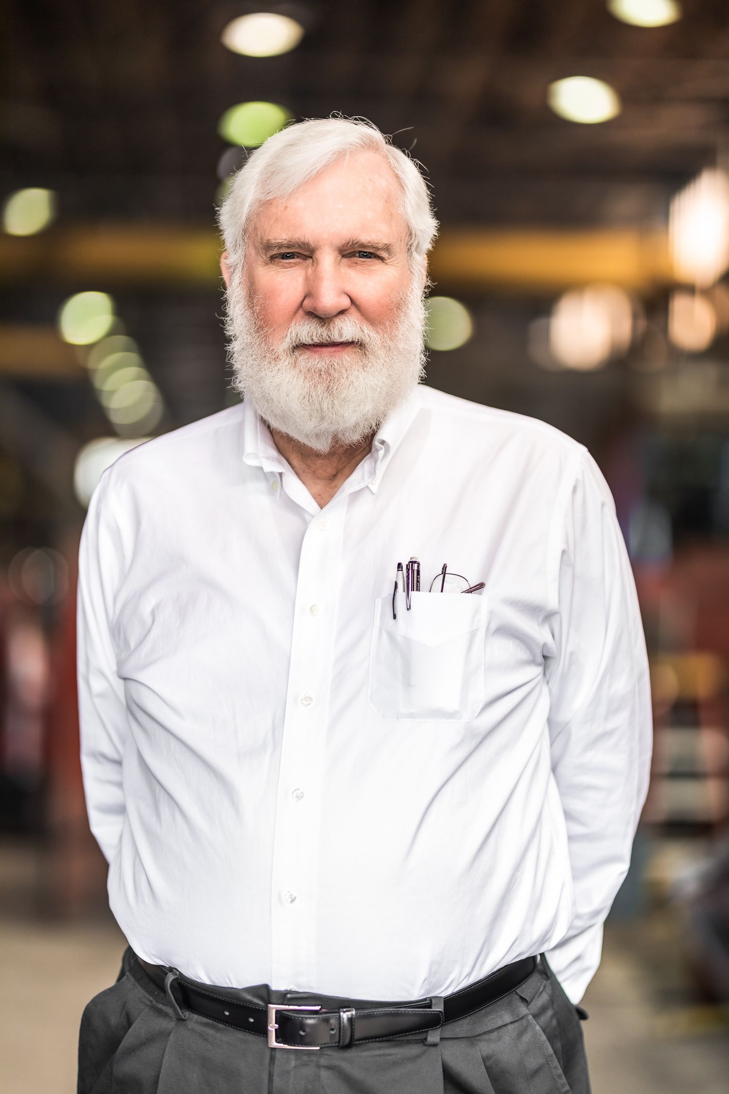 Senior man with white beard and hair in white dress shirt stands in an industrial setting.