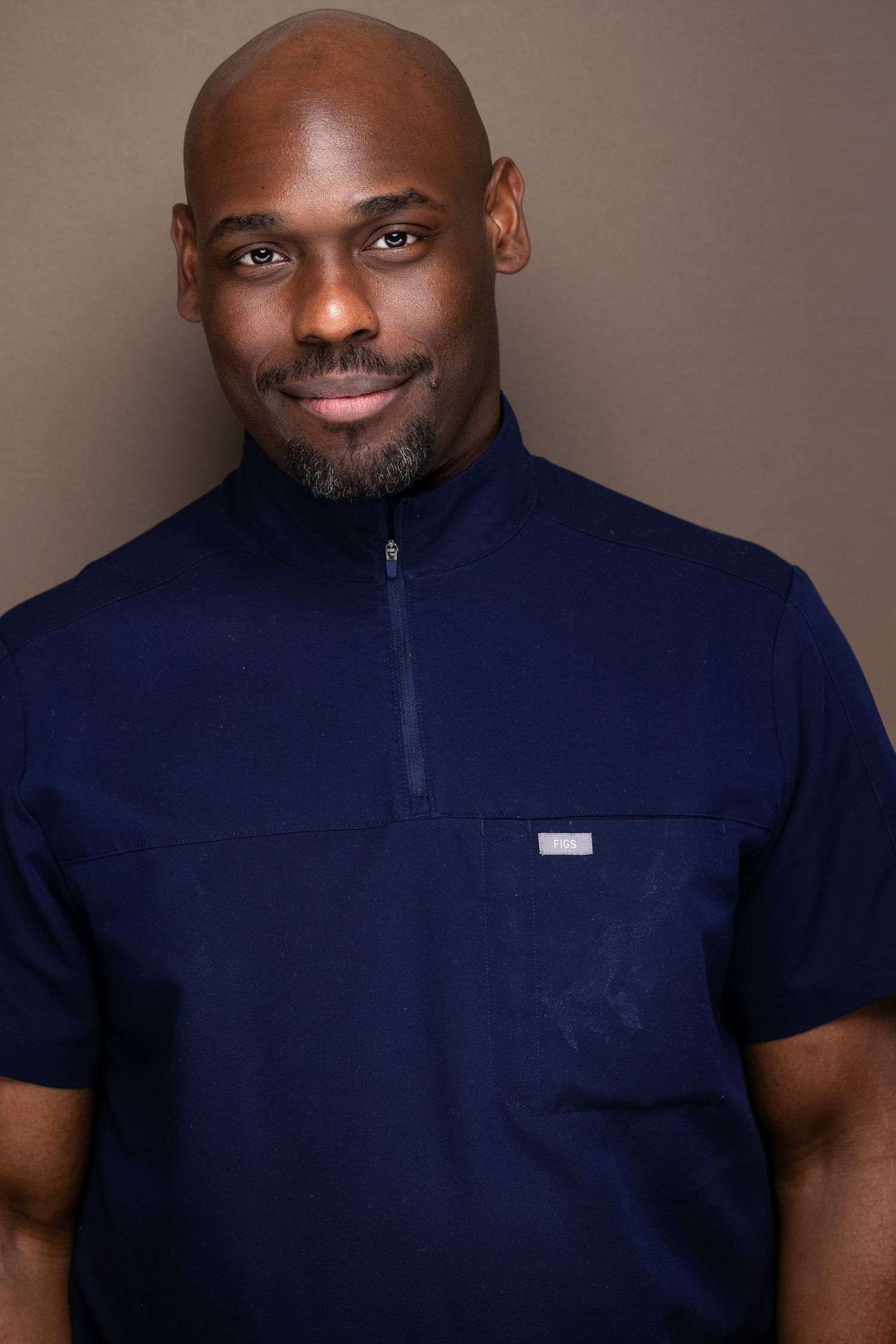 Professional headshot of a bald man with a goatee wearing a navy zip-collar short-sleeve shirt against gray backdrop.