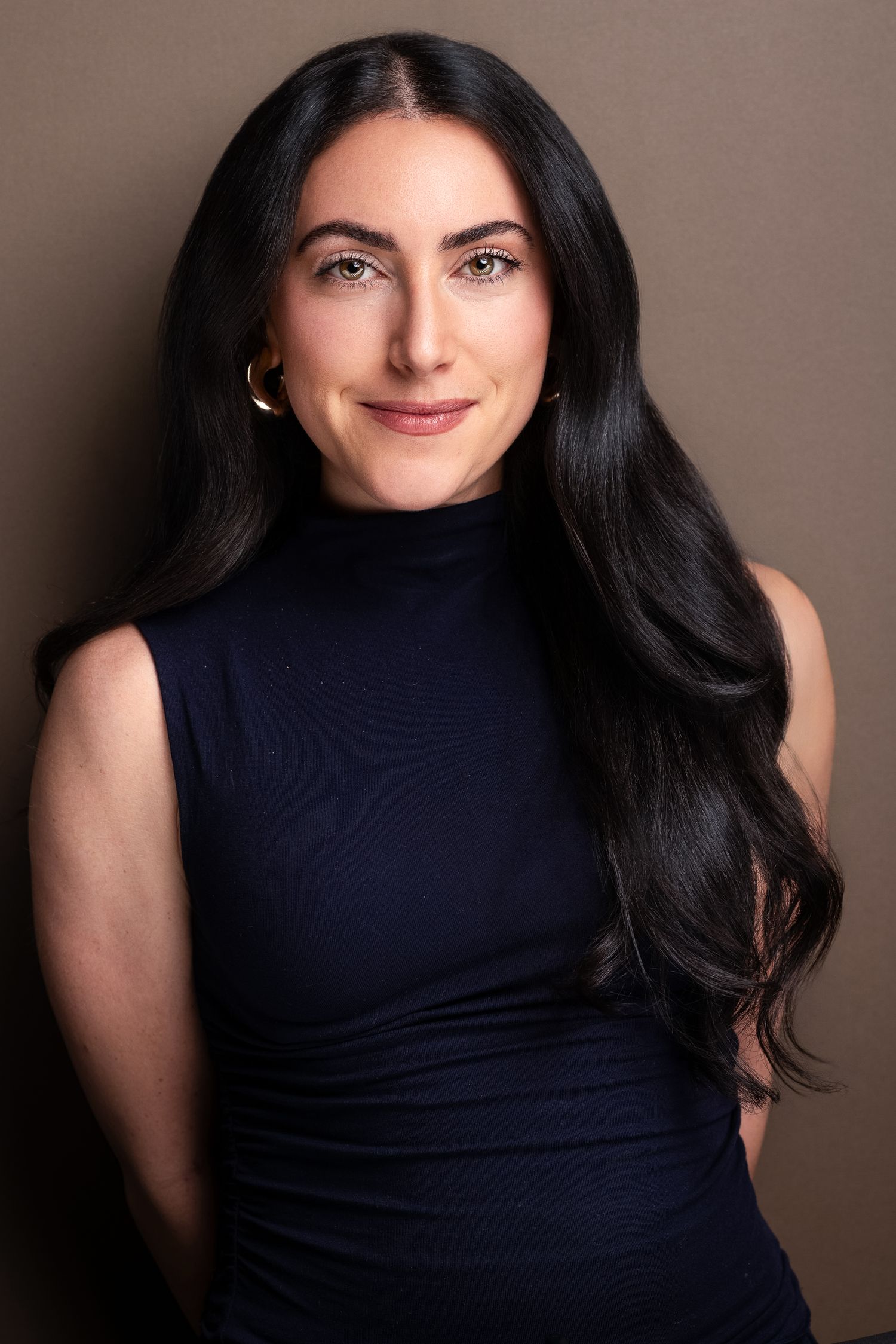 Woman with long black hair wearing a navy turtleneck top and gold hoop earrings poses for a professional headshot.