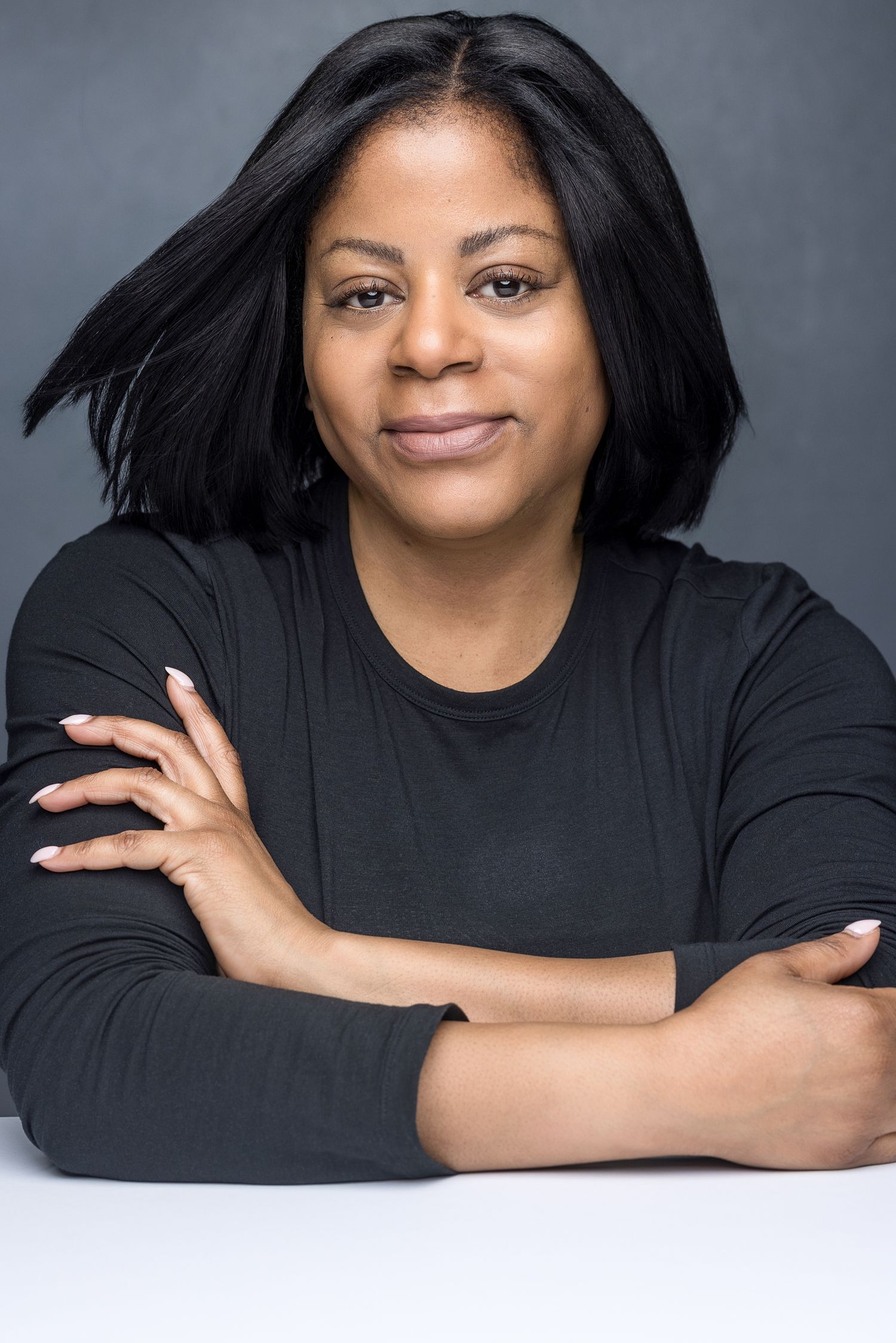 Professional headshot of a confident woman with black hair, arms crossed, wearing a black top against a gray background.