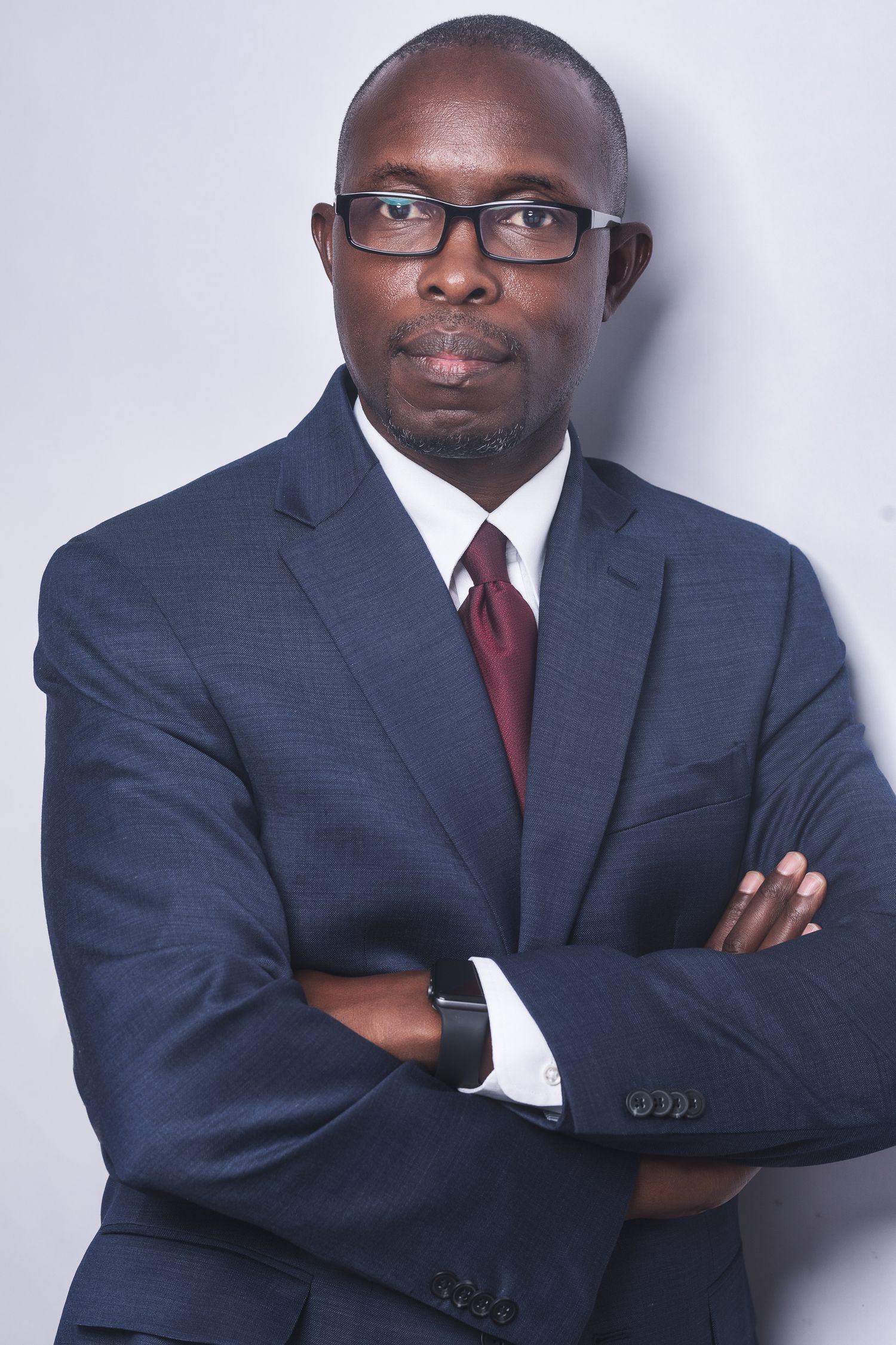 Professional Black man in navy suit and red tie, arms crossed, wearing glasses, confident business portrait.